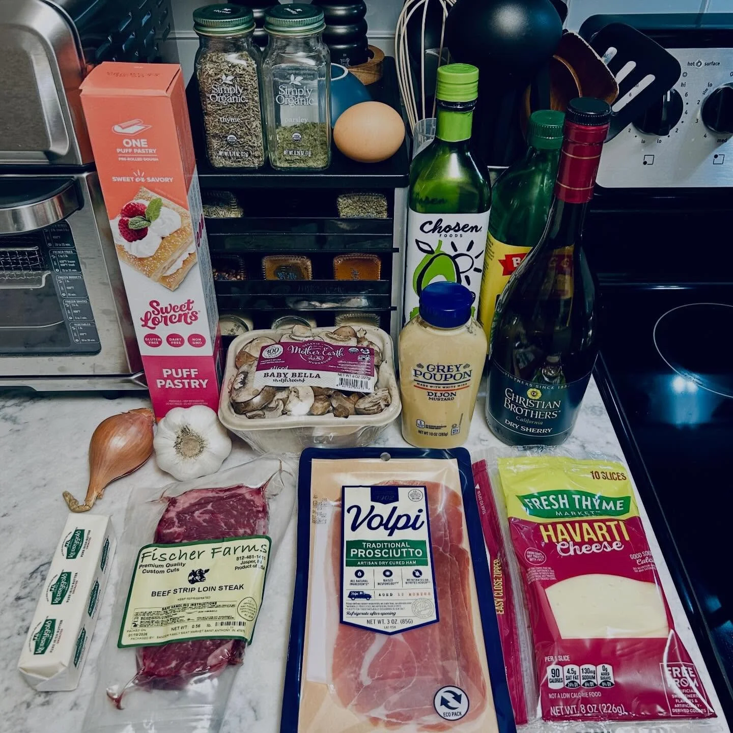 Ingredients for cooking: puff pastry, beef sirloin steak, provolone cheese, Havarti cheese, mushroom, garlic, onion, mustard, sherry wine, dried mushrooms, and various herbs and spices. The background shows kitchen items like spice jars, an egg, and kitchen appliances.