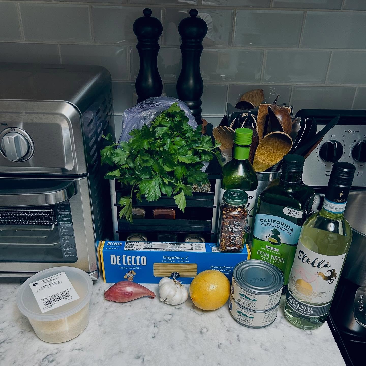 Counter with various ingredients and kitchen items including a bunch of parsley, garlic, lemon, canned fish, olive oil, pasta, spices, and utensils.