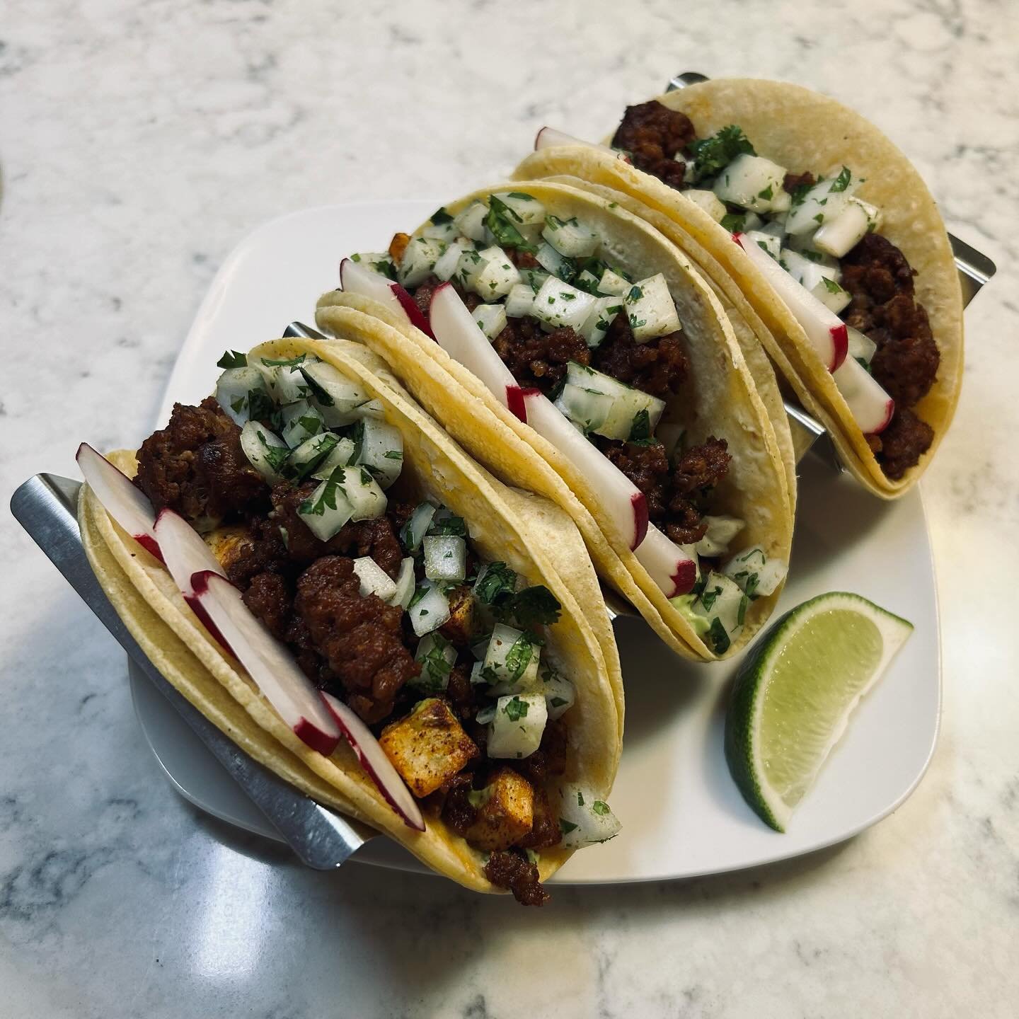 Three tacos filled with seasoned meat, topped with chopped onions and cilantro, served on a white plate with lime wedges, on a marble countertop.
