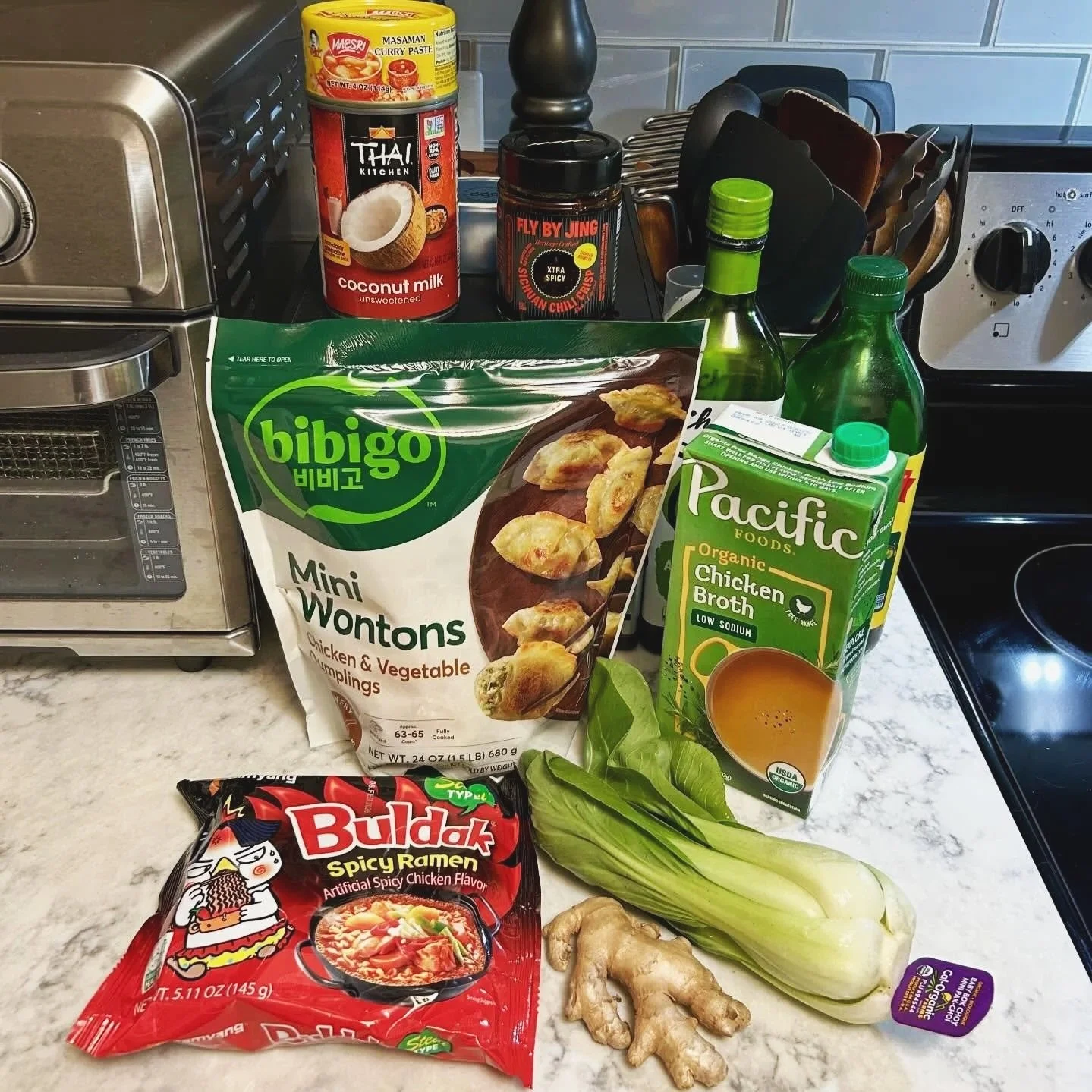 Various Asian cooking ingredients on a kitchen counter, including bibigo mini wontons, Pacific chicken broth, buldak spicy ramen, bok choy, ginger, and condiments.