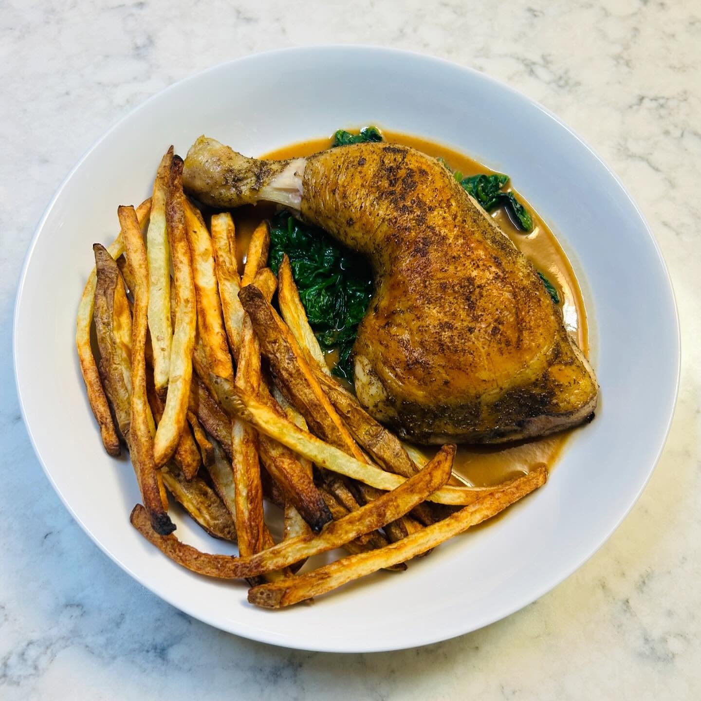 Plate of roasted chicken thigh with crispy French fries and sautéed spinach, served in a light brown sauce.