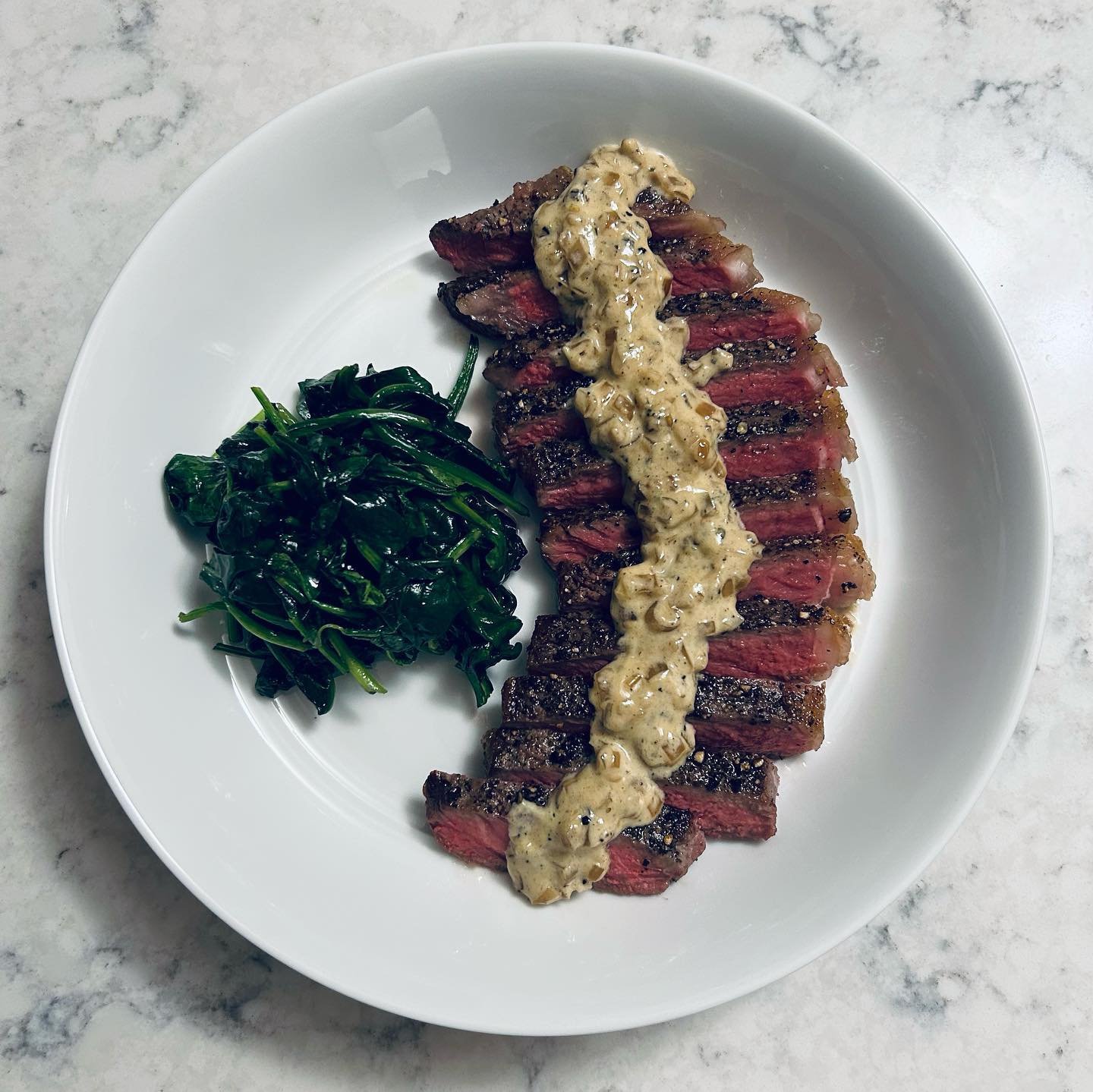 A white plate with sliced medium-rare steak with creamy mushroom sauce and cooked greens on a marble surface.