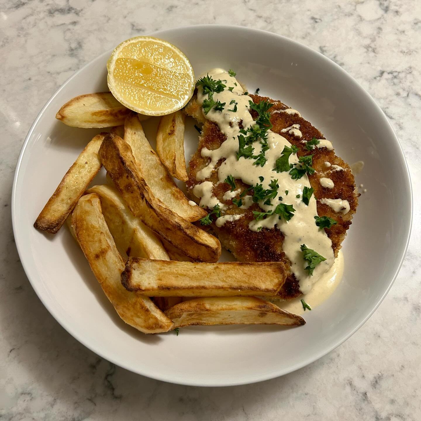 Fried fish fillet topped with white sauce and chopped parsley, served with lemon wedge and French fries on a white plate