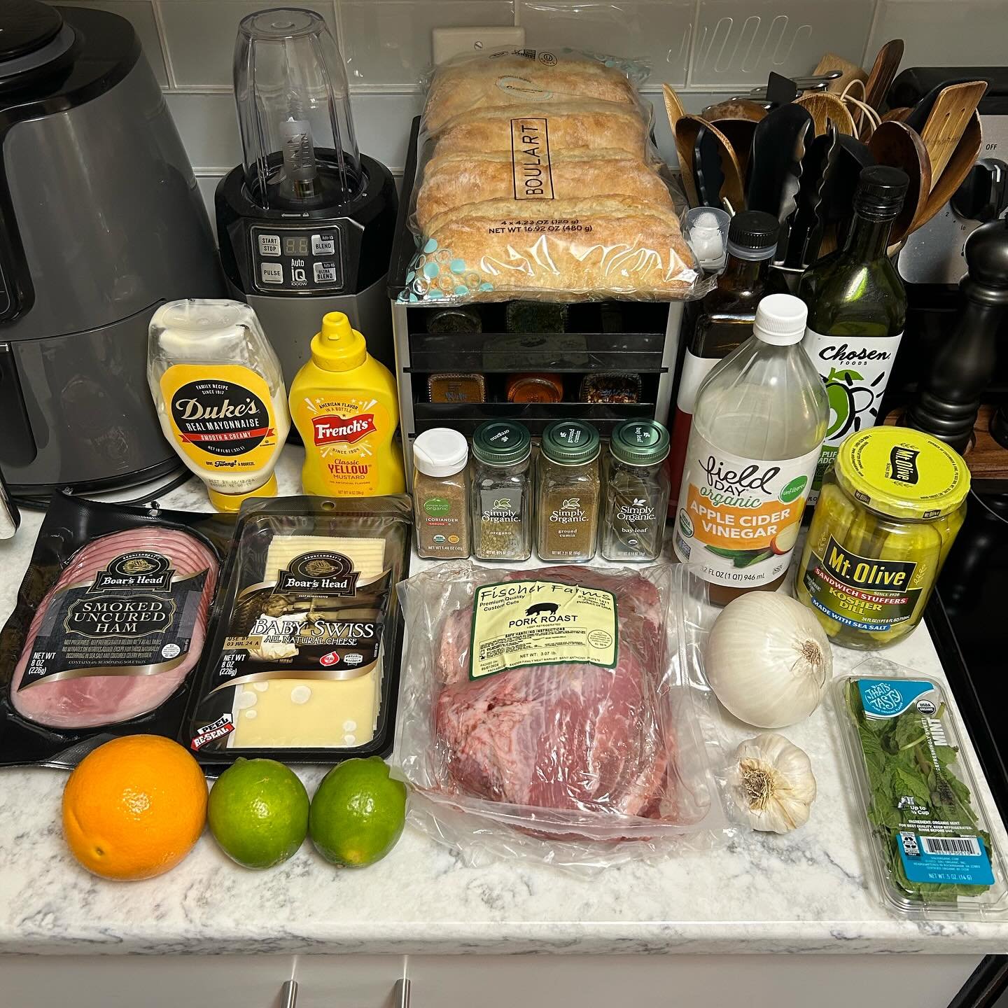 Counter with bread, ham, cheese, pork roast, garlic, citrus fruits, herbs, condiments, vinegar, olive jar, and various kitchen items.