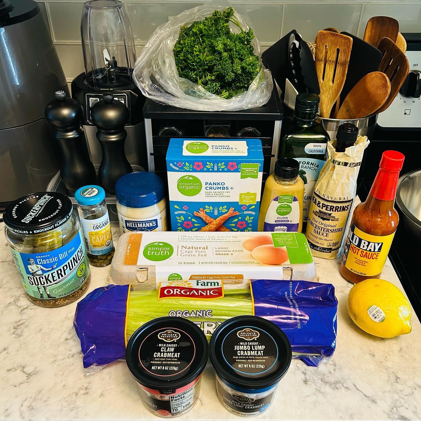 Collection of groceries on a kitchen countertop, including eggs, lemon, crabmeat, hot sauce, pickles, seasonings, and condiments.