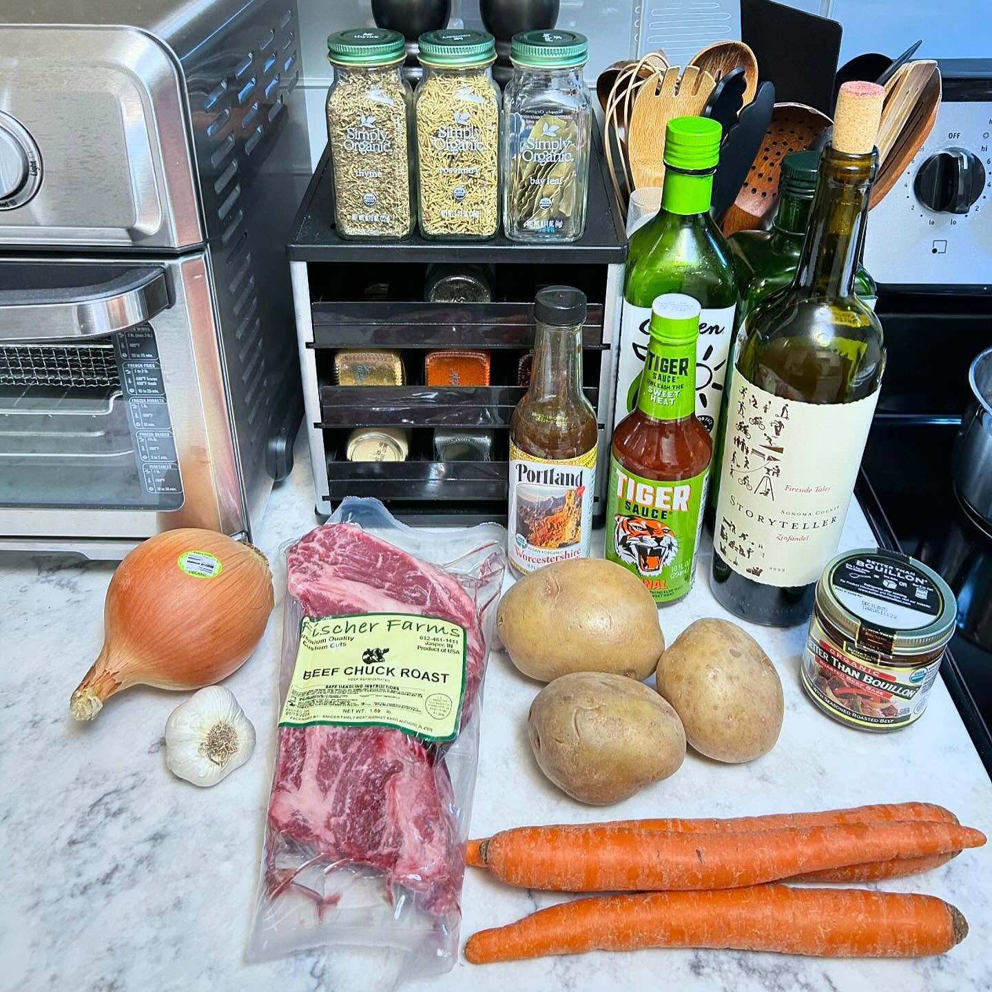 On a kitchen counter, there are fresh vegetables including two carrots, three potatoes, an onion, and a head of garlic. There is also a package of beef chuck roast, bottles of Worcestershire sauce, Tiger sauce, a bottle of wine, and a jar of bouillon. In the background, there are spice jars, wooden utensils, and an oven.