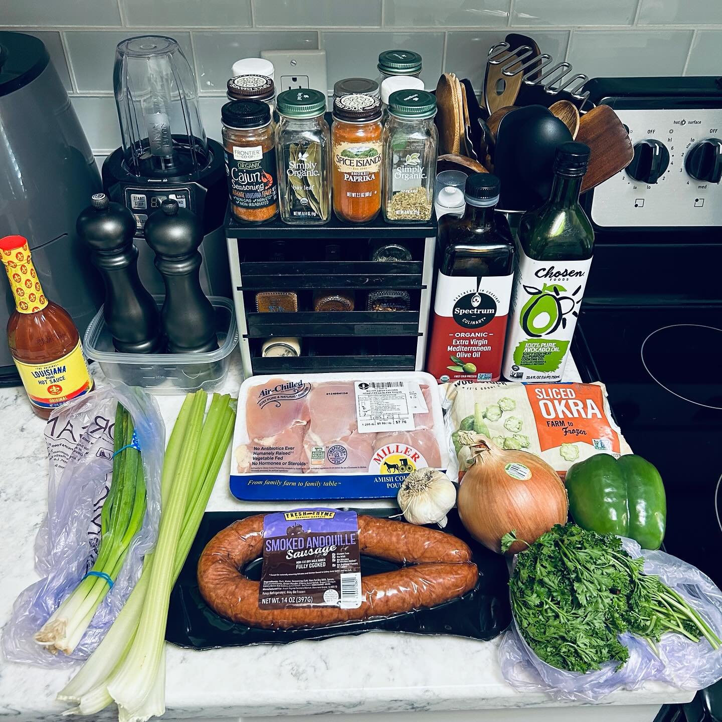 Fresh ingredients including celery stalks, smoked andouille sausage, chicken thighs, garlic, onion, green bell pepper, parsley, and canned condiments on a kitchen countertop, with spices and oil in the background.