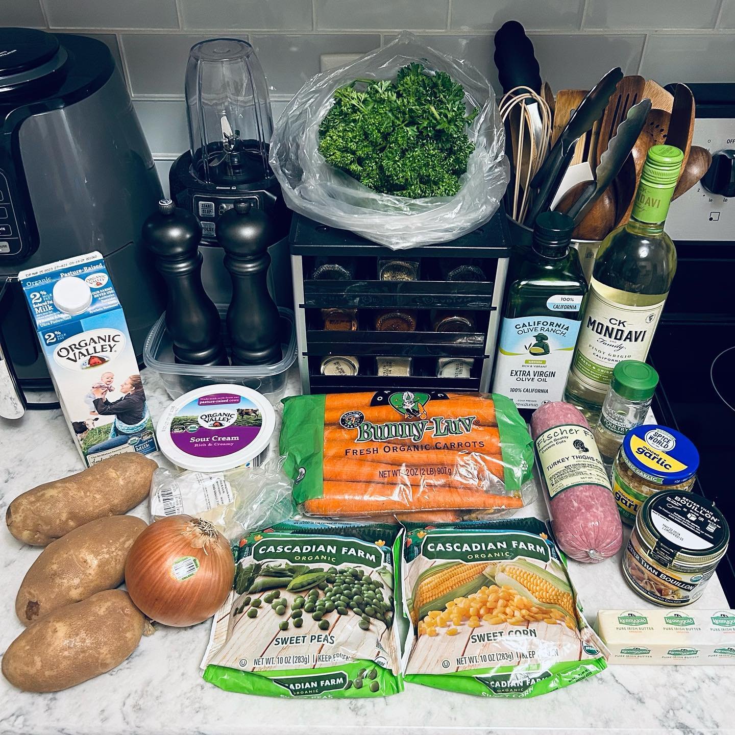 A variety of fresh groceries on a kitchen counter including potatoes, an onion, sweet peas, sweet corn, a package of carrots, sour cream, chicken thighs, olive oil, garlic, seasoning, and a bunch of parsley.
