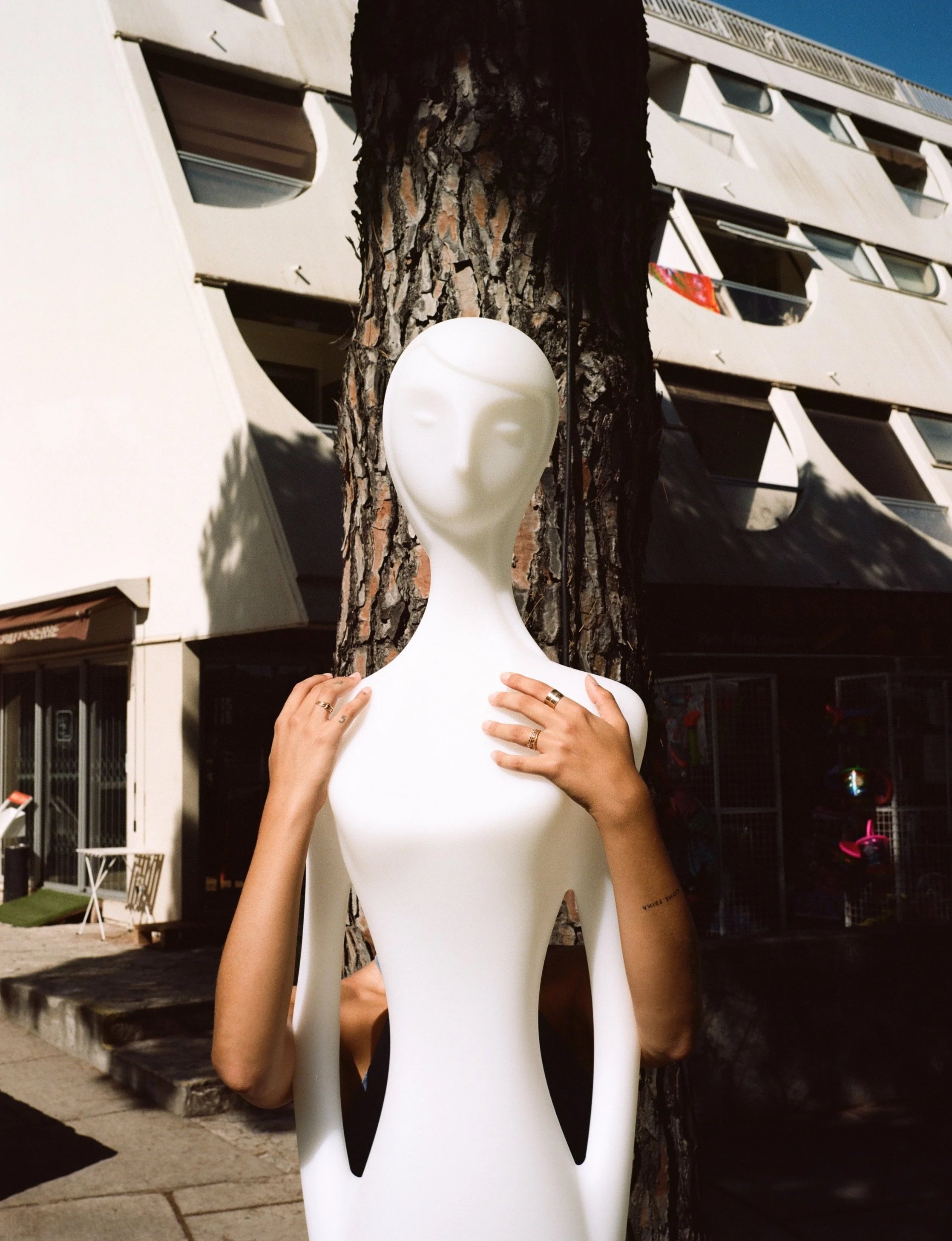A white mannequin with a smooth, featureless face is standing outdoors behind a tree, with a person partially obscured by the mannequin. The person's hands rest on the mannequin's shoulders, and they are wearing rings and a tattoo. Background shows a modern building with balconies and windows.