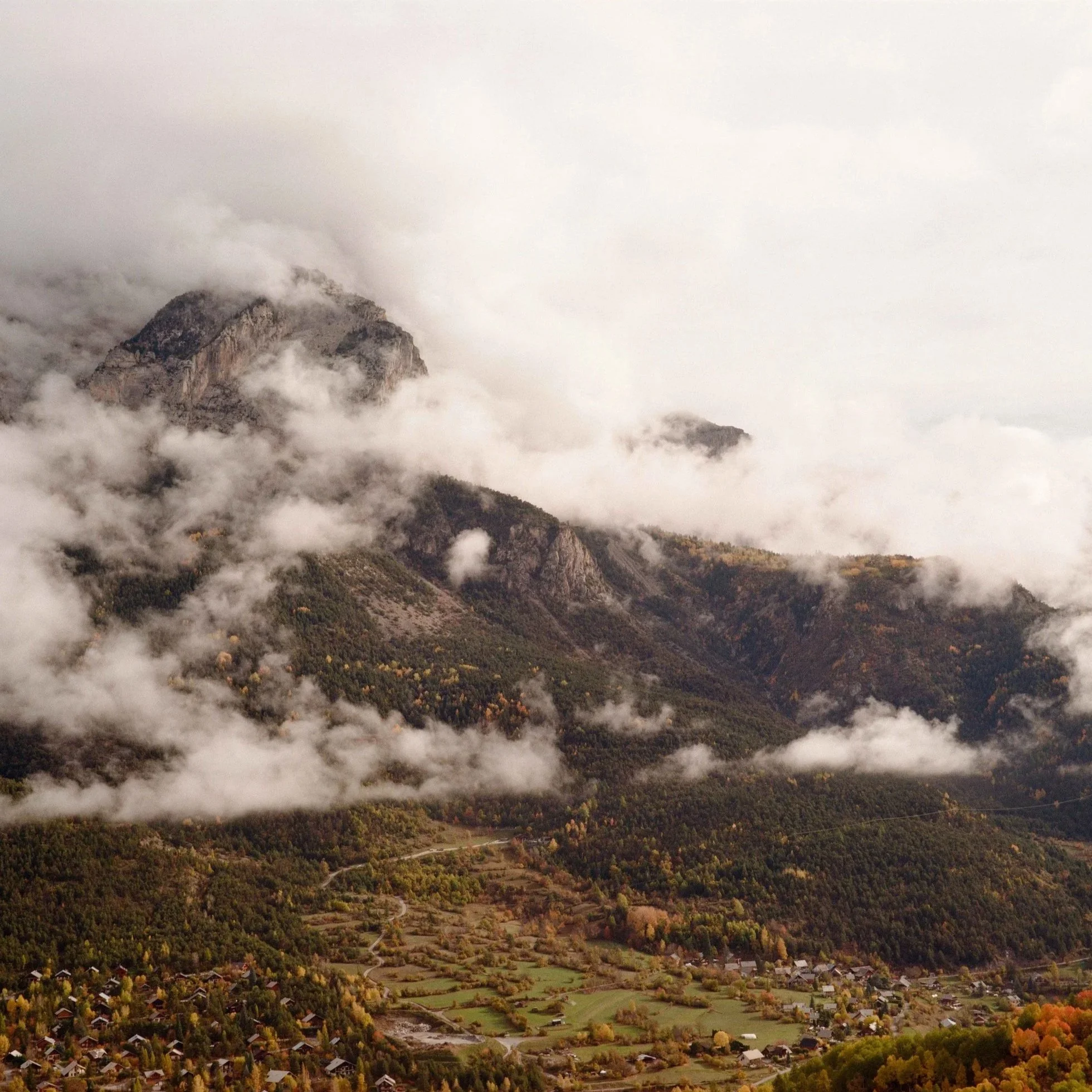 Mountain landscape with peaks covered in clouds and a valley with scattered houses and autumn foliage.
