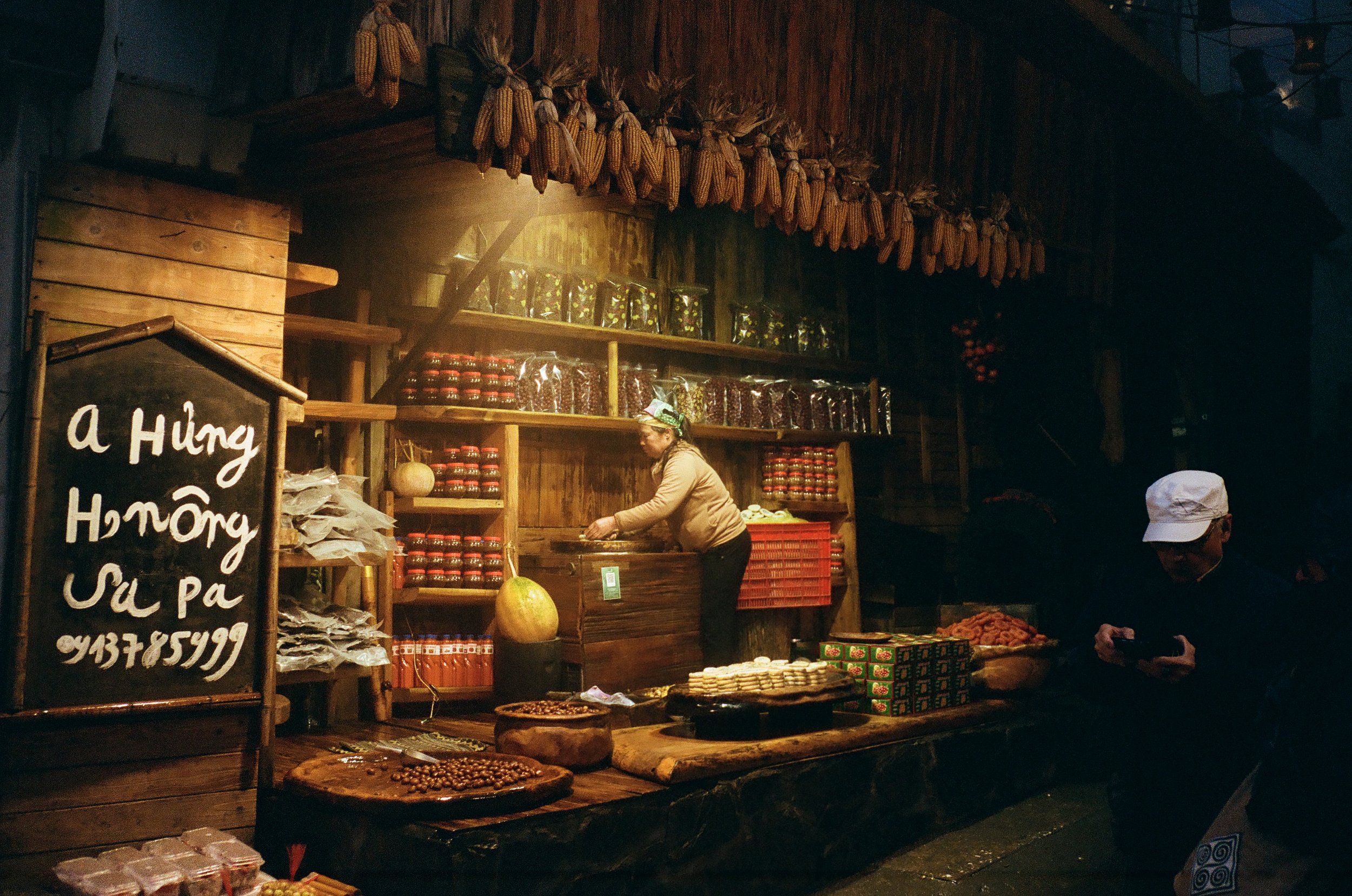 A woman sells jars and sachets of products at an outdoor market stall with a wooden structure. Some products are displayed on shelves and the stall is decorated with hanging ears of dried corn. A man in a white cap is seen looking at his phone nearby.