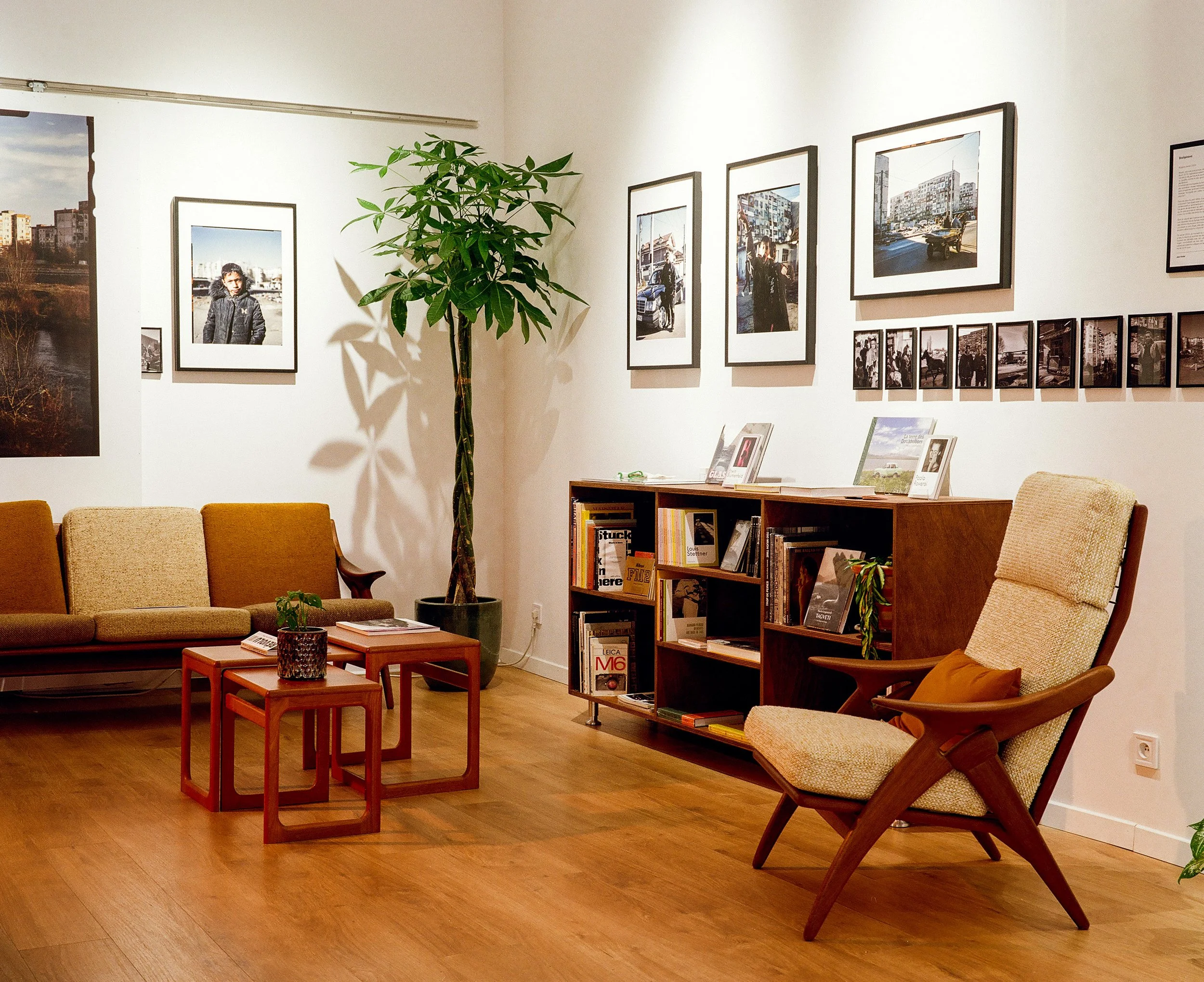 A cozy living room with a beige armchair, a tan sofa, a wooden shelf filled with books and photos, a potted plant, framed photographs on the white walls, and small wooden tables with decorative items. The flooring is hardwood.