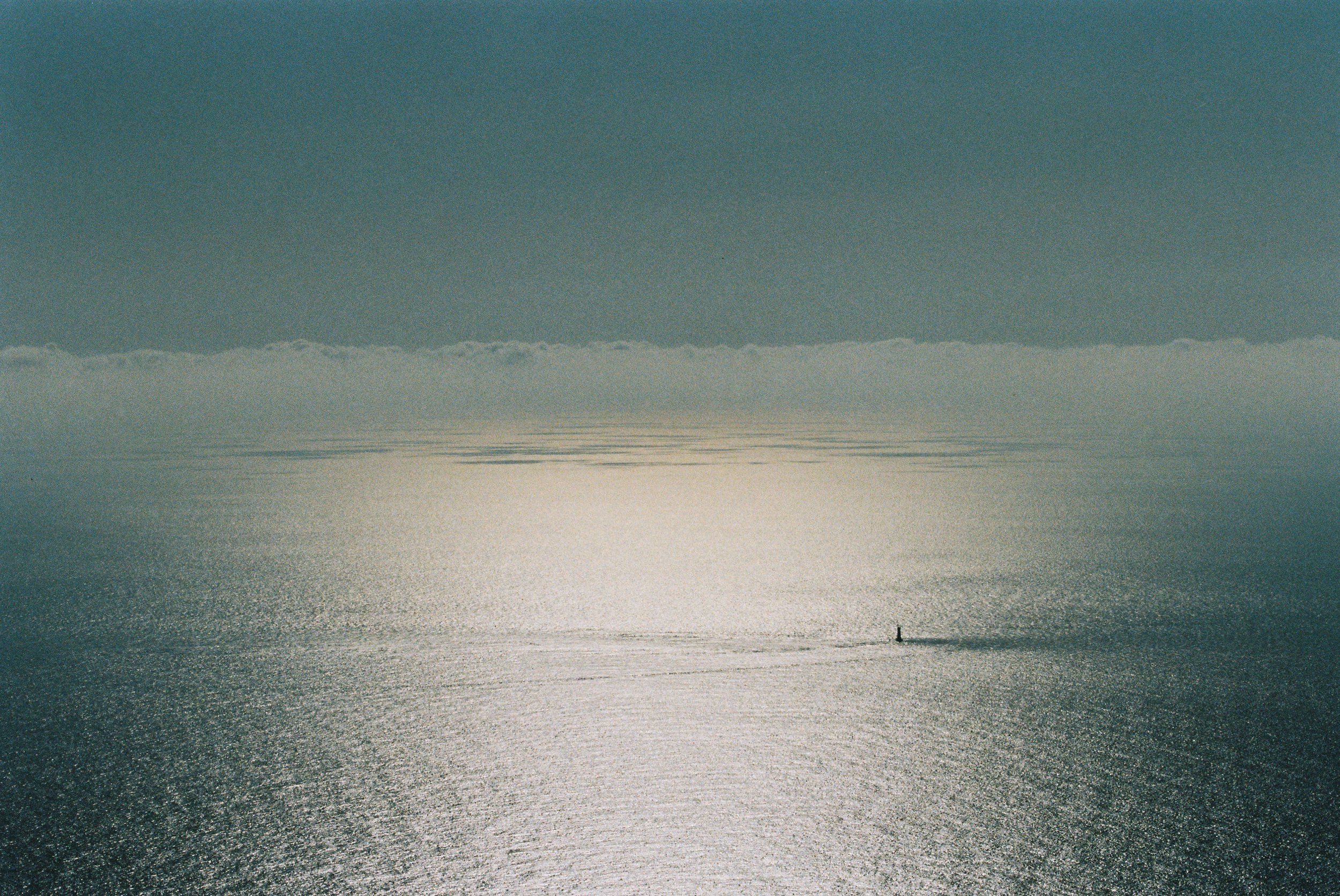 A boat on a calm body of water with a person standing on it, reflecting sunlight, and a distant shoreline with clouds in the sky.