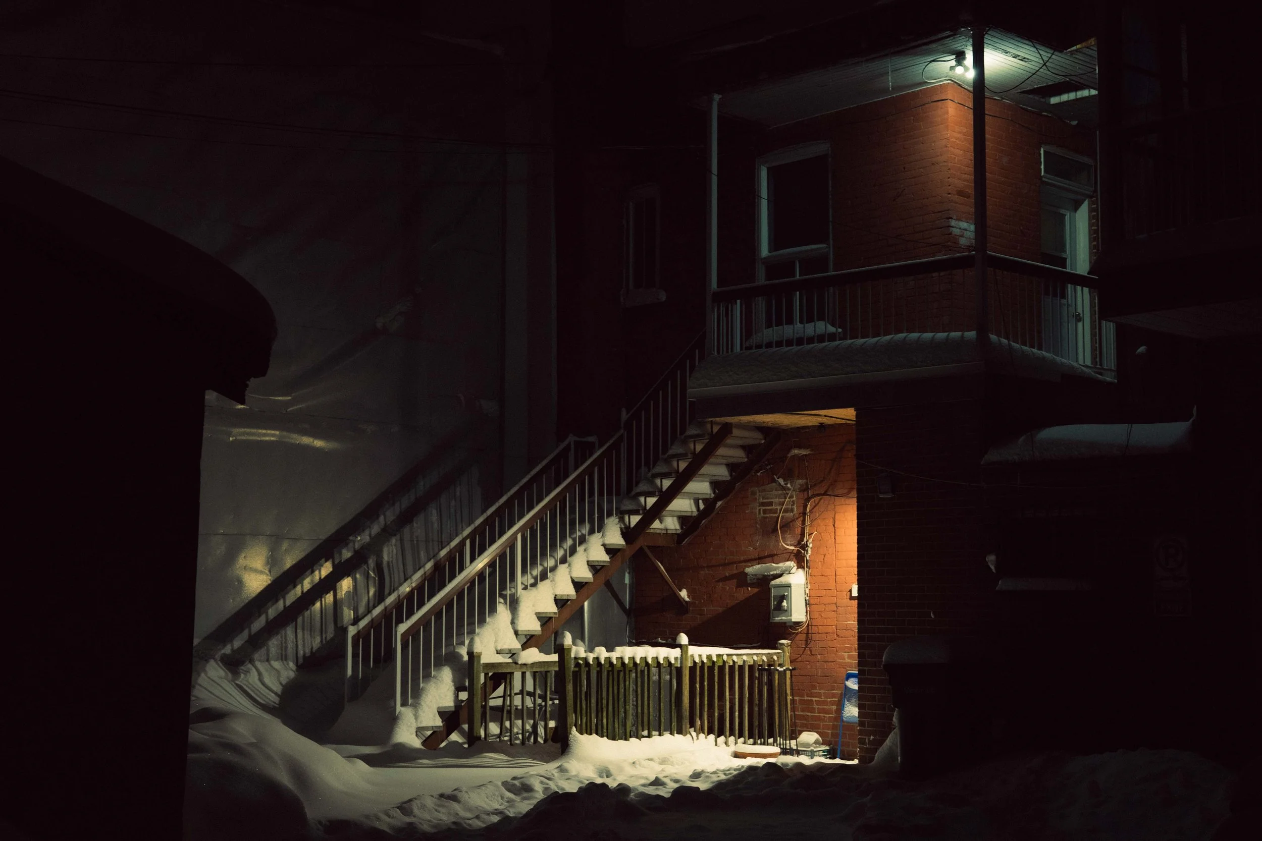 Nighttime scene of a brick apartment building with an outdoor staircase covered in snow, illuminated by a nearby light, with snow accumulated on the ground and stairs.