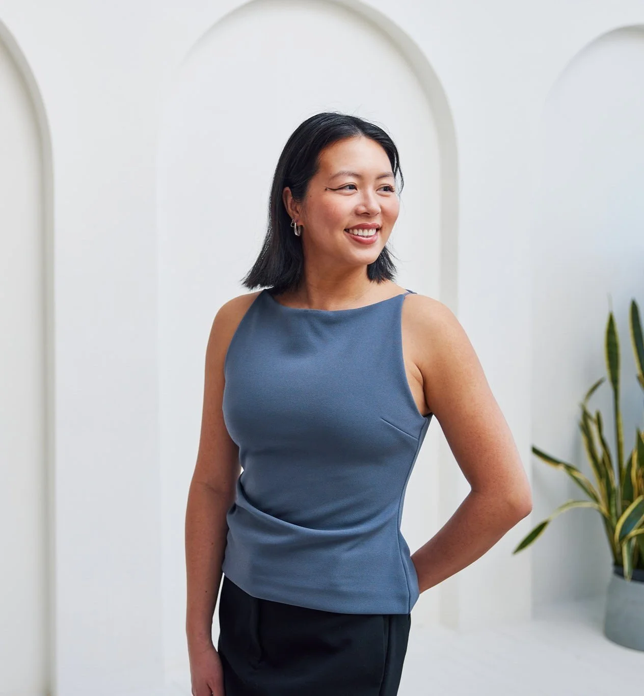 A woman with black hair smiling, wearing a blue sleeveless top and black pants, standing indoors near white walls and a potted plant.