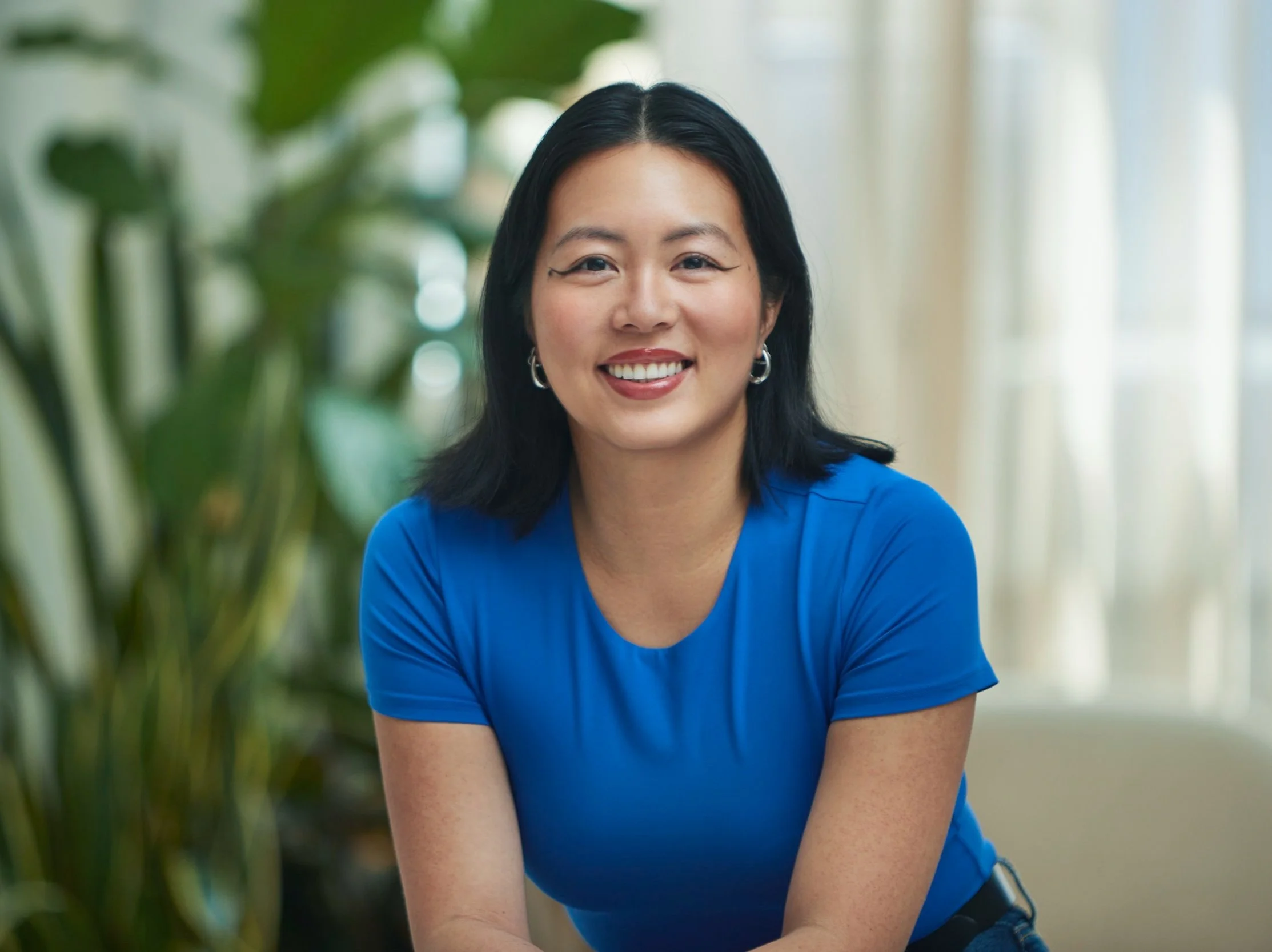 A smiling woman in a bright blue top sitting indoors near green plants and large windows