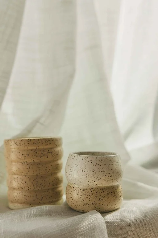 Two speckled ceramic cups with a textured surface, placed on a white fabric surface with a white curtain in the background.