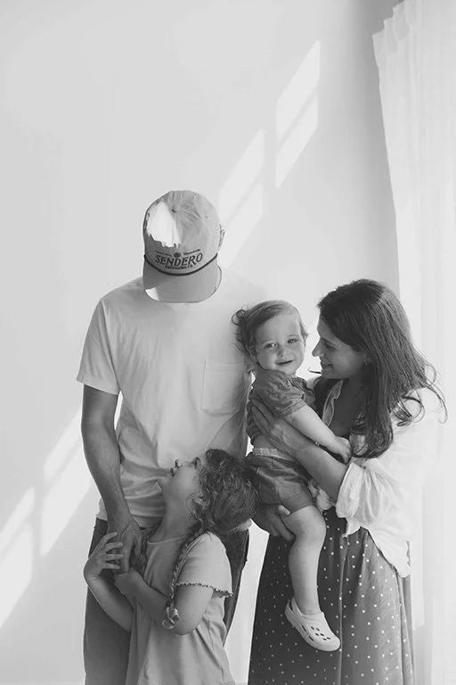 A family of four, including a father, mother, and two young daughters, smiling and enjoying time together indoors, with sunlight streaming through a window.