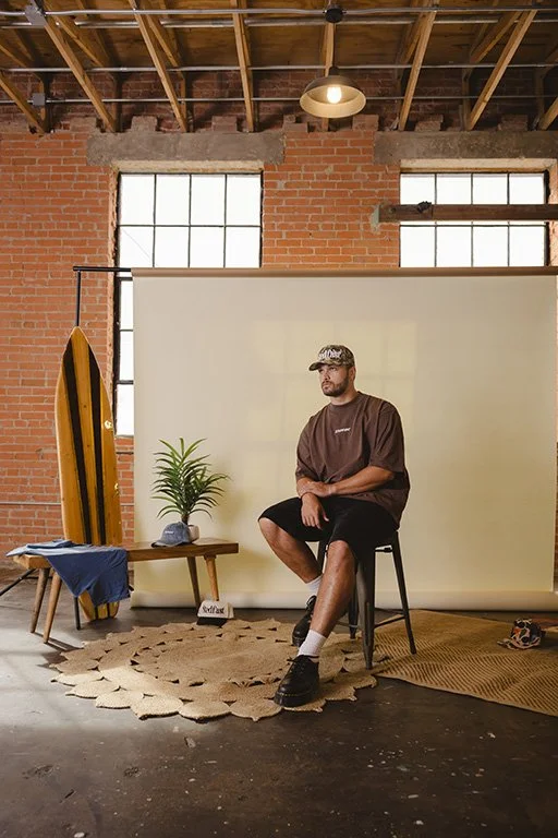 A man sitting on a stool in a studio with a brick wall, large windows, a potted plant, surfboards, and a backdrop. The floor has a woven rug and a small table with clothing and a hat.