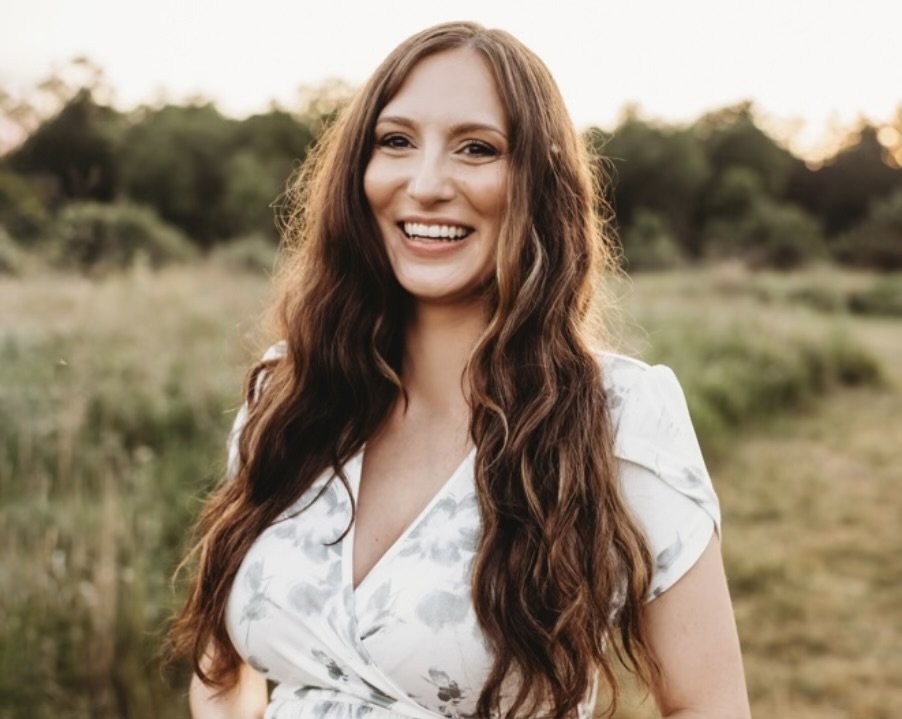 An inviting woman who is a therapist with long, wavy brown hair smiling outdoors in a natural setting with trees and grass at sunset.