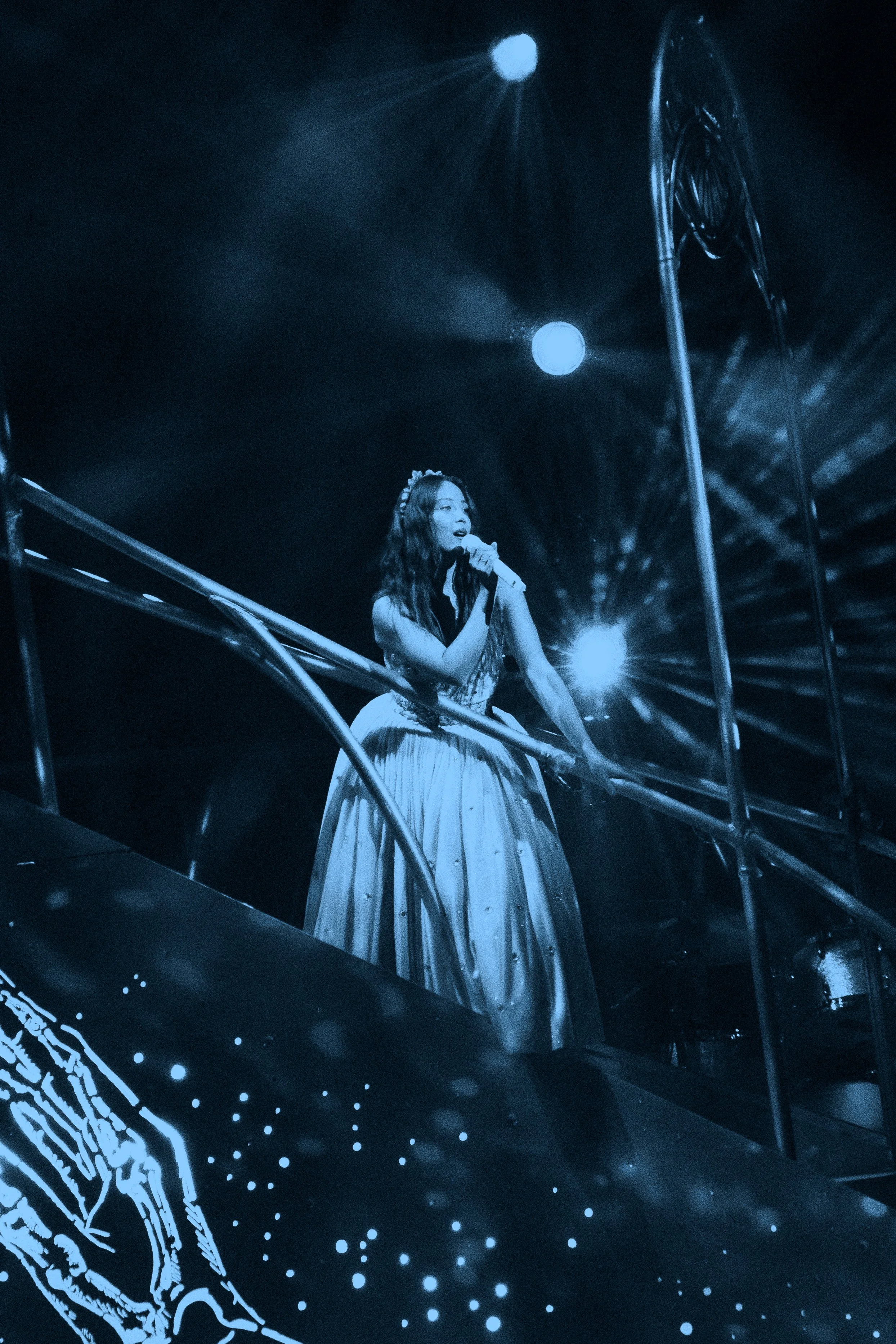 Laufey in a long dress sings into a microphone on a stage with dramatic blue lighting and star-like effects.