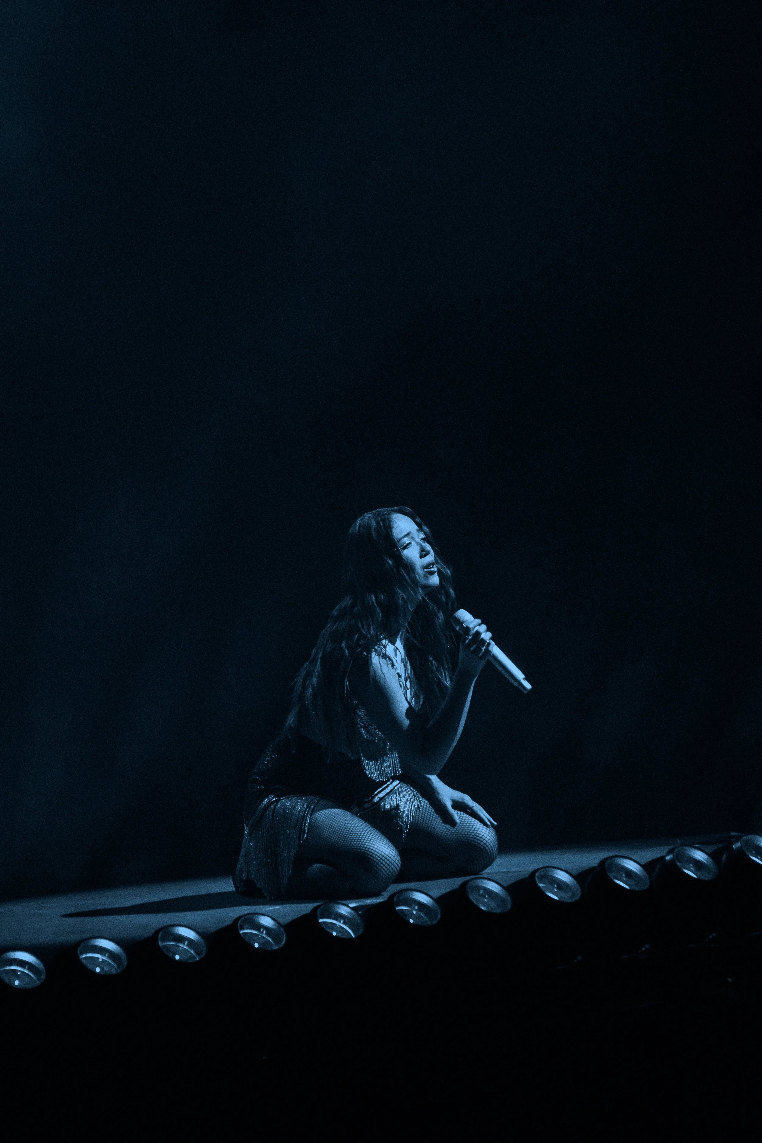 Laufey kneeling on stage, holding a microphone, illuminated in blue light, with a dark background and stage lights in front.