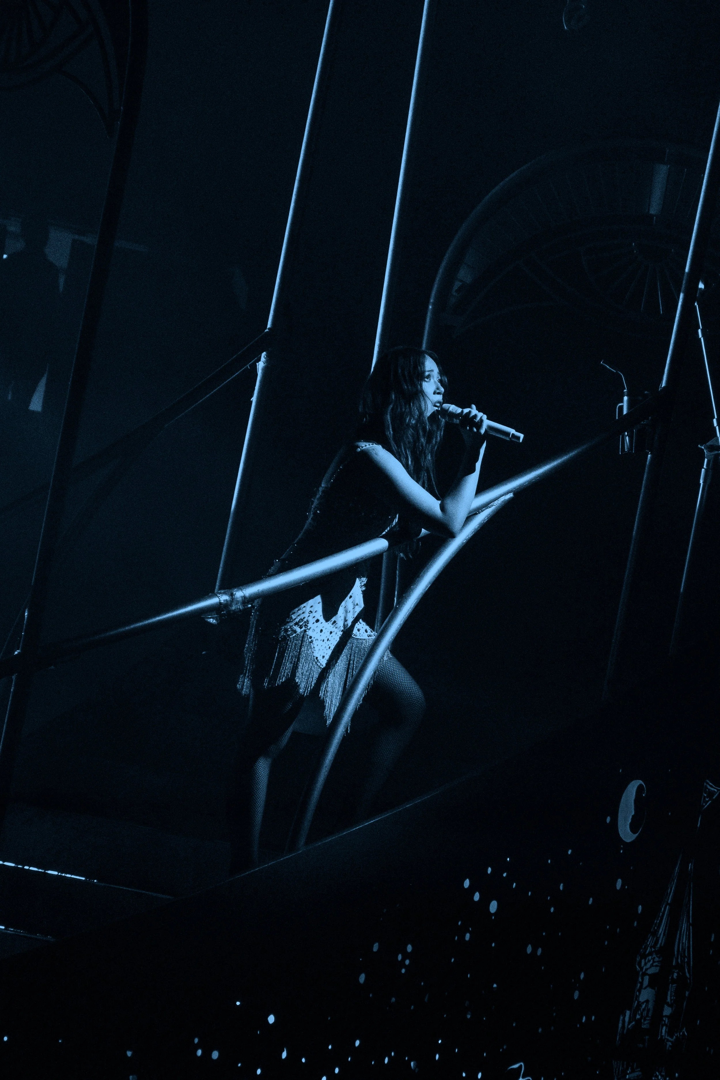 Laufey performing on a dark stage, holding a microphone, with a spiral staircase and minimal lighting.
