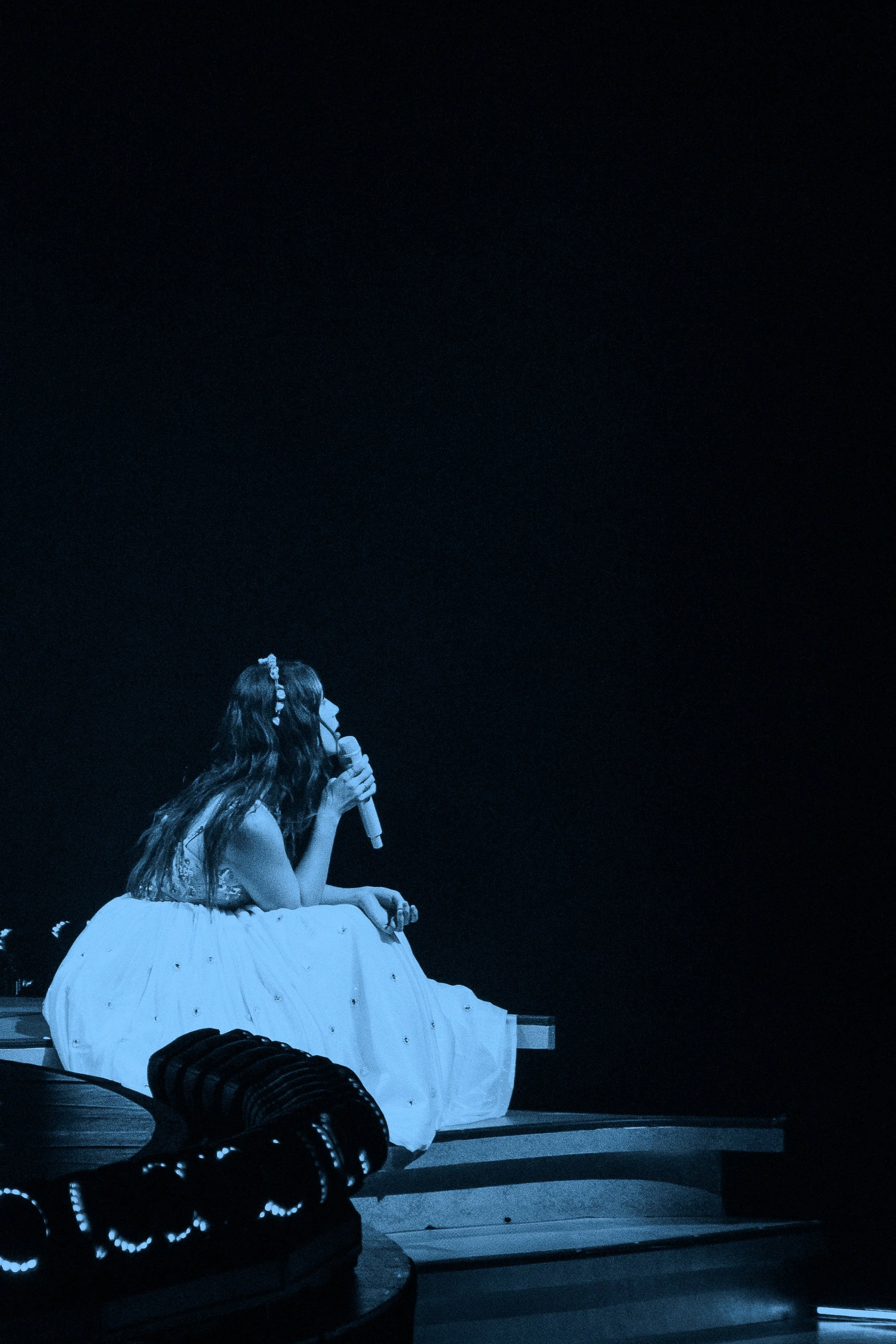 Laufey sitting on a bench at night, holding a microphone near her mouth, dressed in a white skirt and top, with her hair decorated with small flowers.