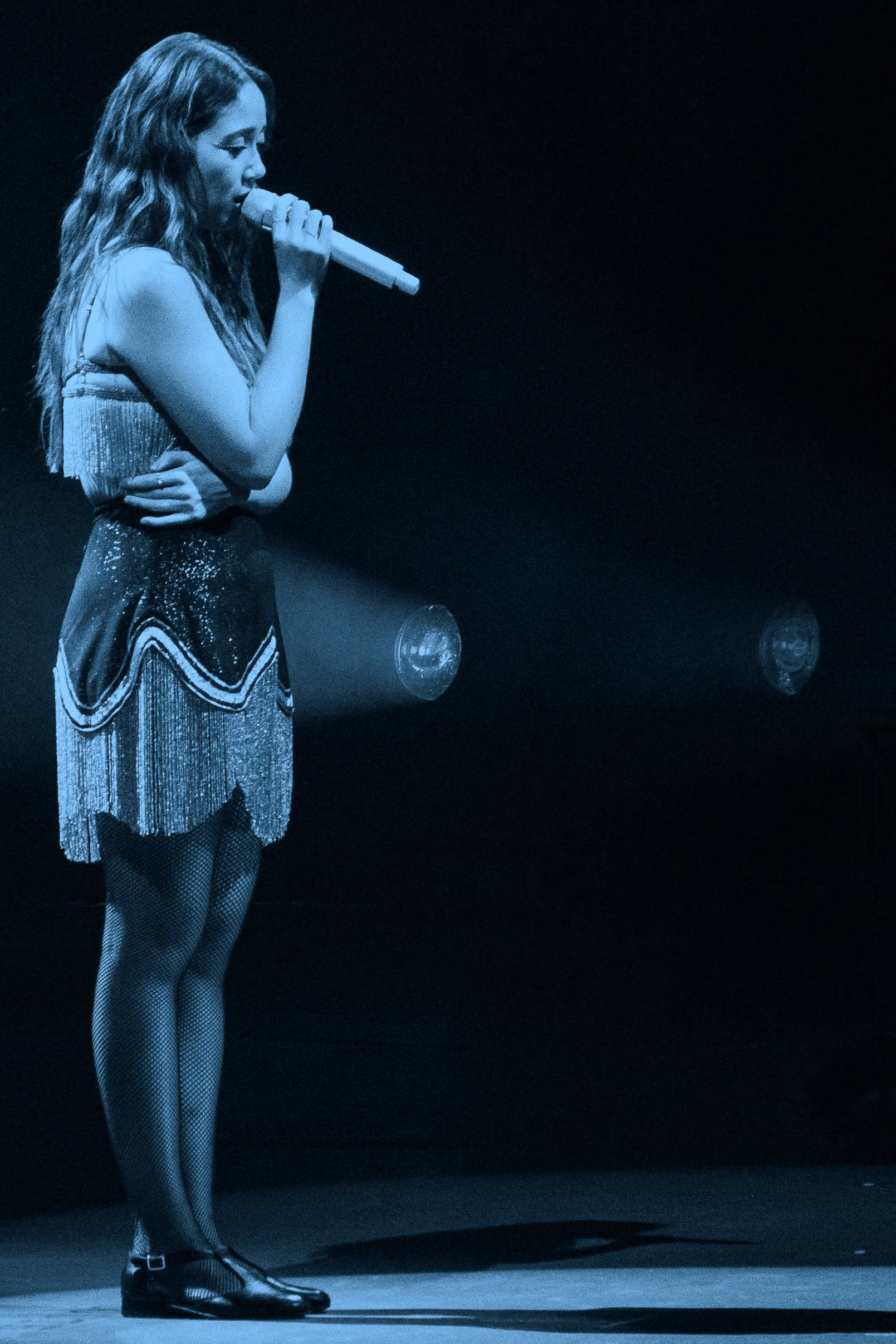 Laufey with wavy hair singing into a microphone on stage, wearing a fringed top, a sequined skirt, fishnet stockings, and black shoes, stage lighting in the background.