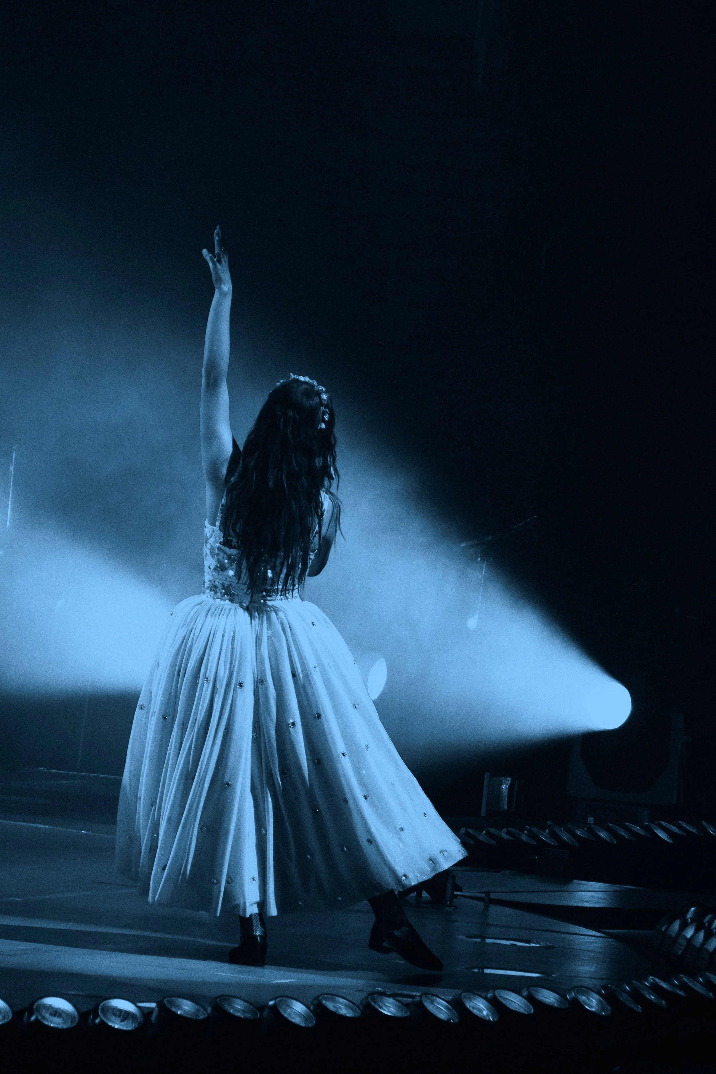 Laufey performing on stage in a white gown with embellishments, illuminated by a spotlight, with smoke and stage equipment in the background.