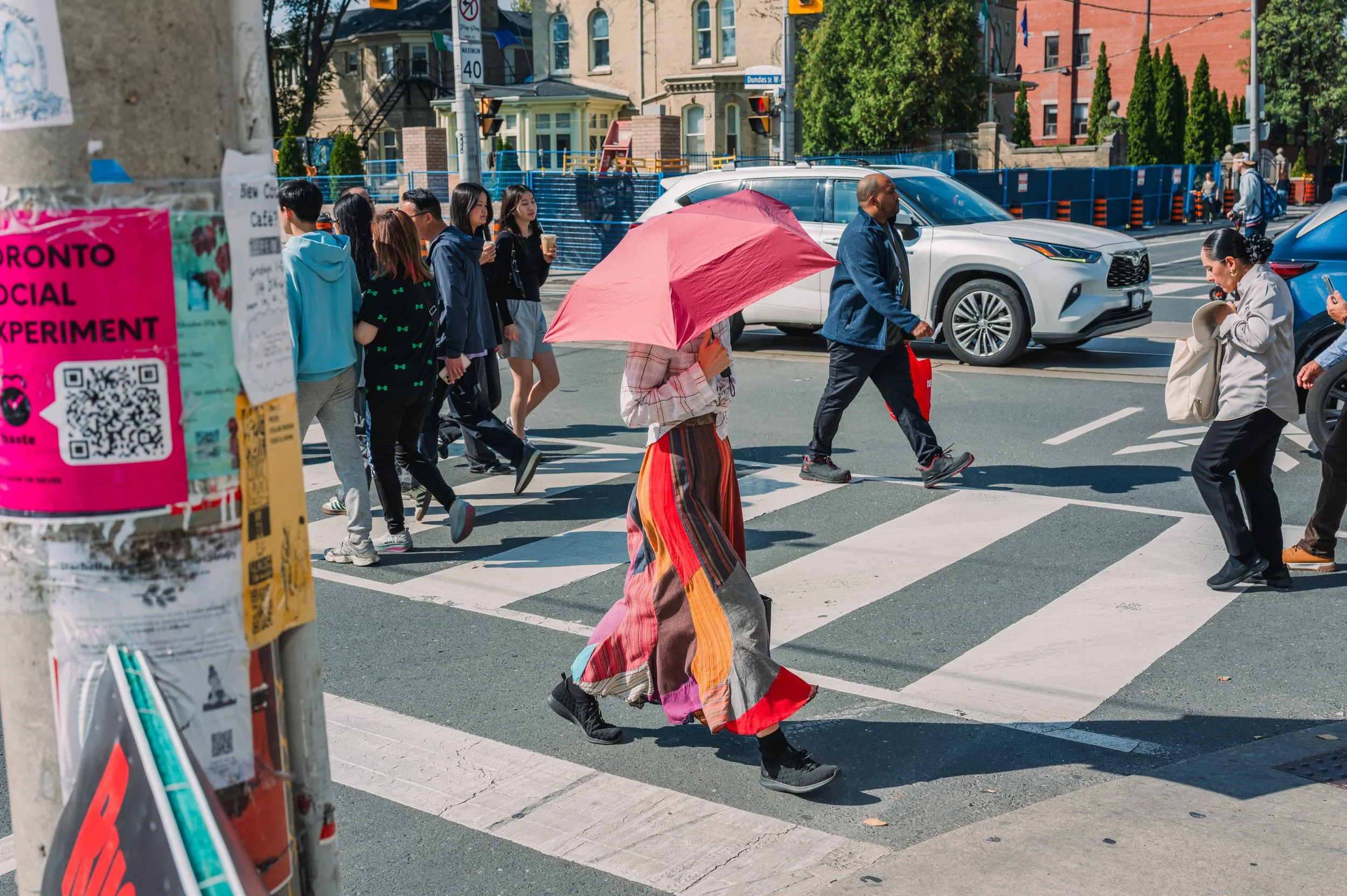 A woman walking across a crosswalk holding a pink umbrella in a busy city street with several pedestrians and cars.