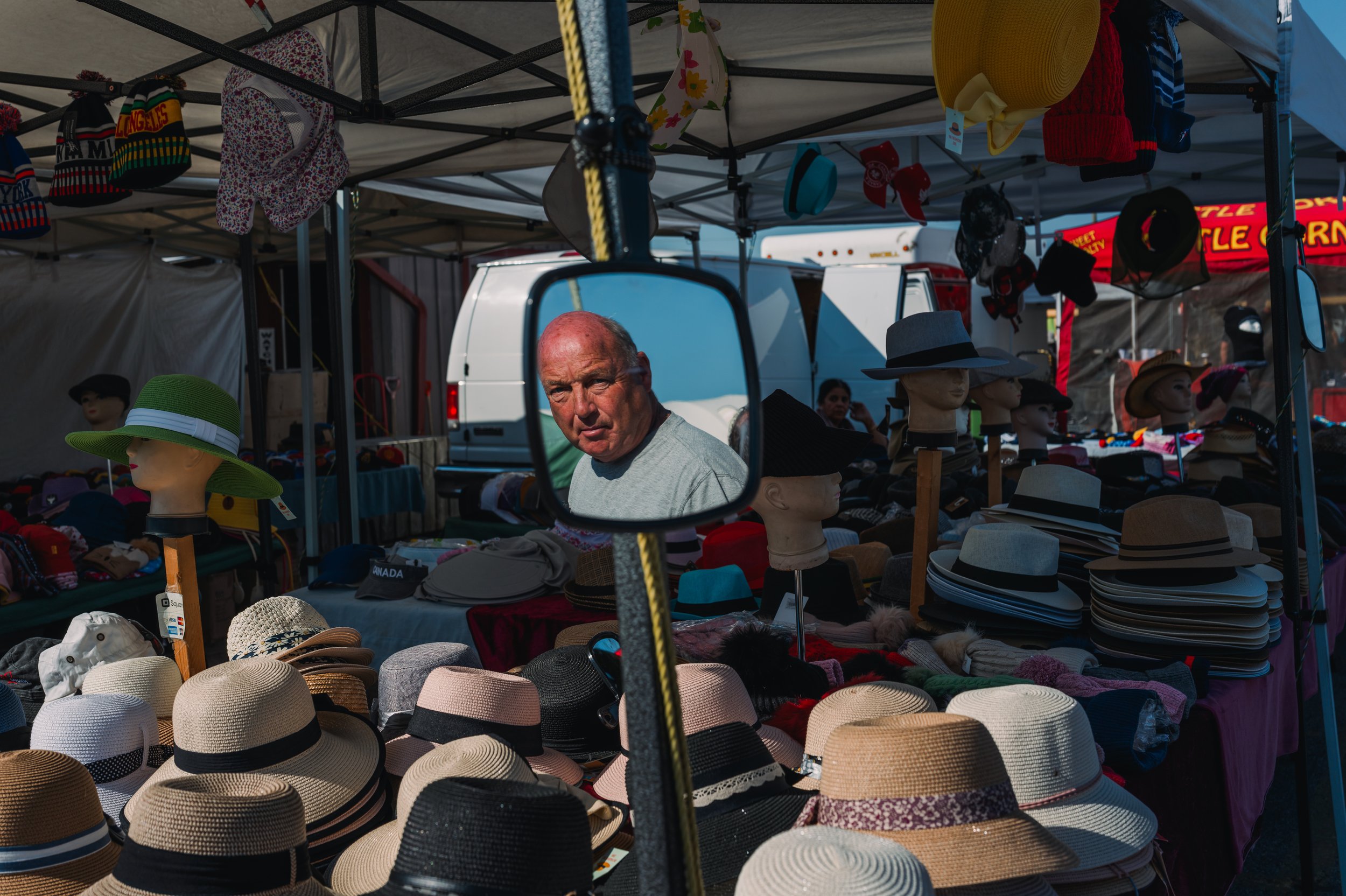 A man with a serious expression looking into a mirror at a hat stall at an outdoor market. The stall displays various hats including wide-brimmed straw hats and many mannequin heads wearing hats. The market is covered with tents and other vendors' bo