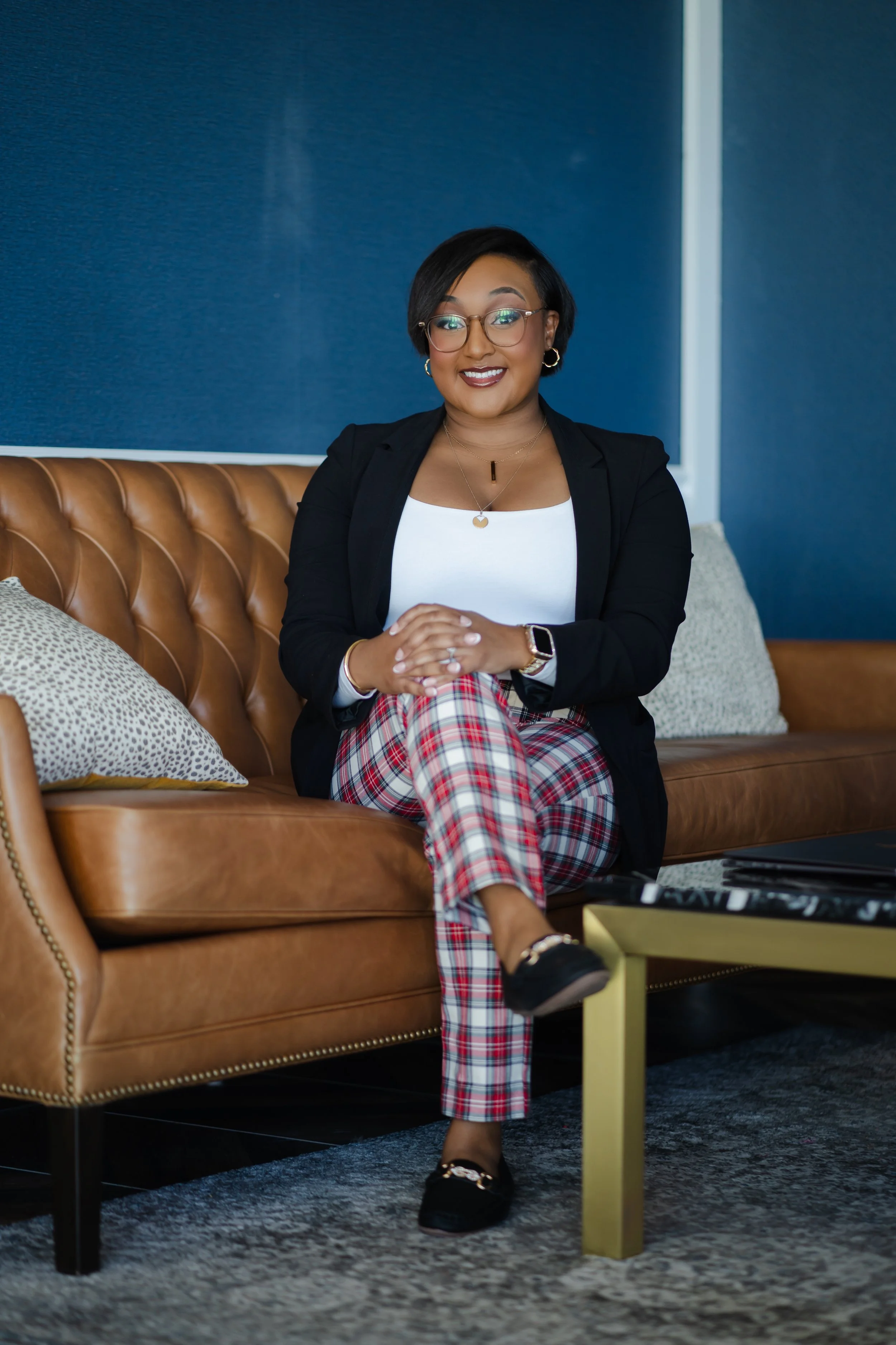 A woman sitting on a tan leather sofa with a navy blue wall behind her, wearing glasses, a black blazer, a white top, plaid pants, and black loafers, smiling with her hands clasped.