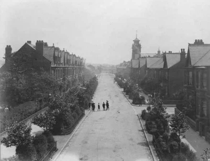 Black and white photo of a wide street in a city with buildings on both sides, trees along the sidewalks, and a few people walking in the middle of the street. A tall clock tower is visible in the background.