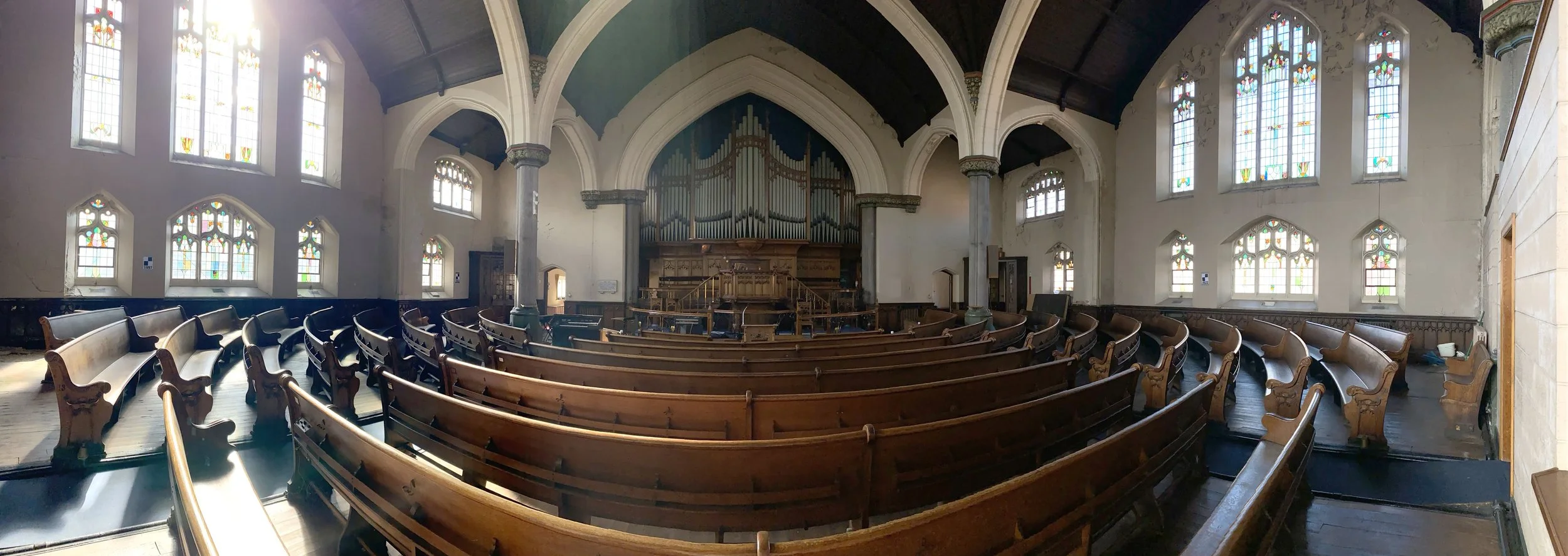 Interior of a historic church with wooden pews, stained glass windows, an organ at the front, and natural light streaming in.
