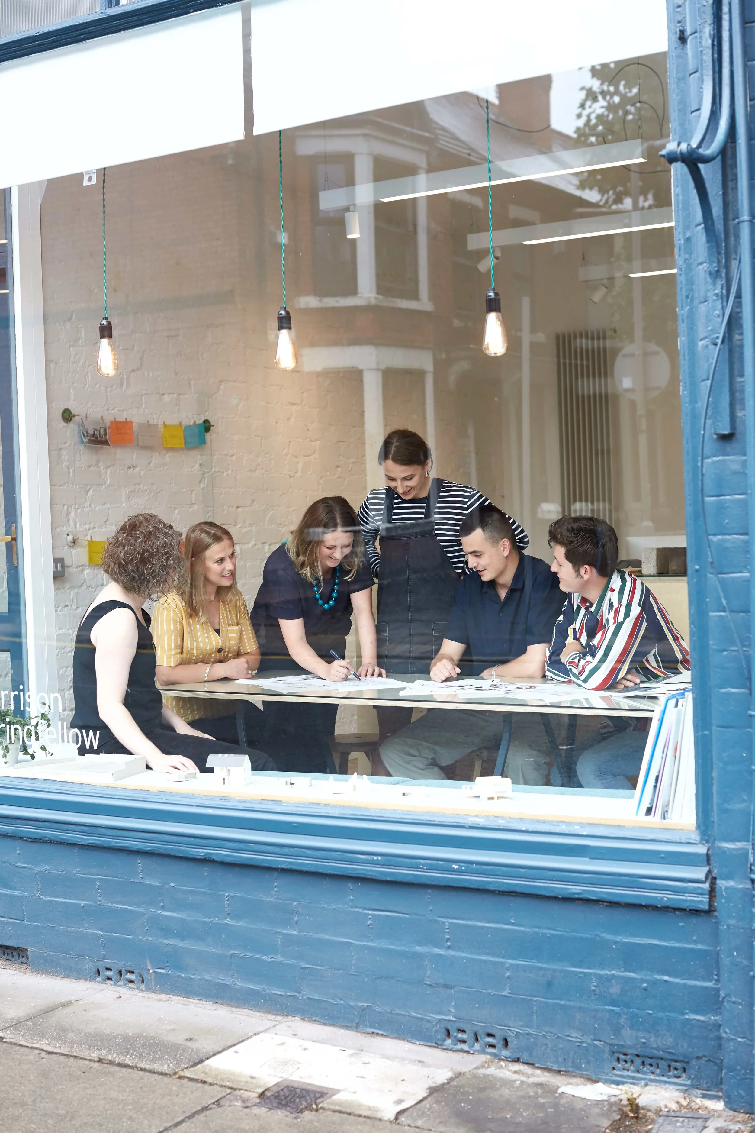A group of six young adults in a meeting room, with five seated around a table and one standing, all looking at papers and enjoying a discussion.