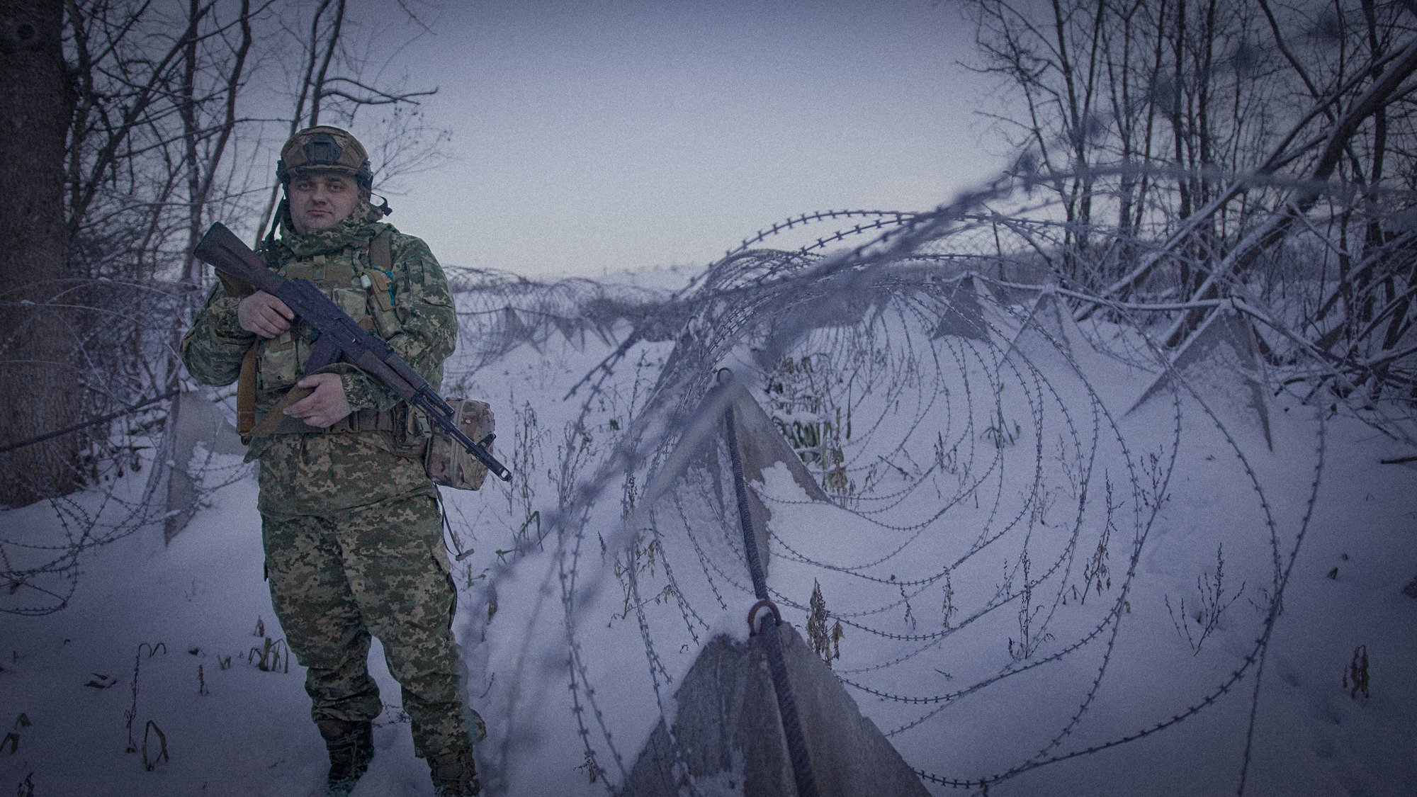 A soldier in camouflage uniform and helmet standing in a snowy wooded area next to barbed wire fence with snow and leafless trees in the background.