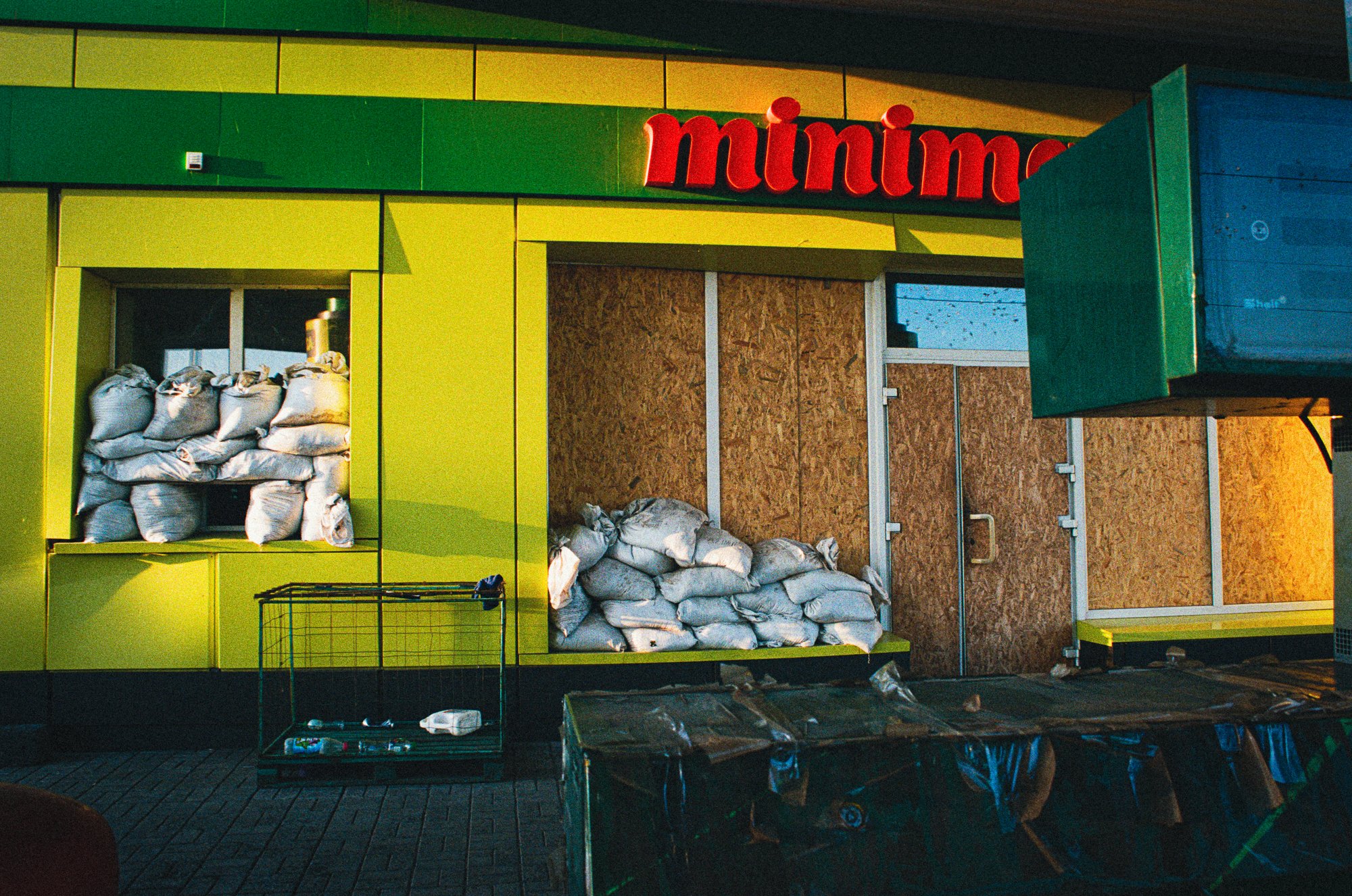 Part of a storefront with green and yellow panels, a red sign with the word 'minimo', and bags of supplies stacked inside and outside, with some items in carts and on the ground.