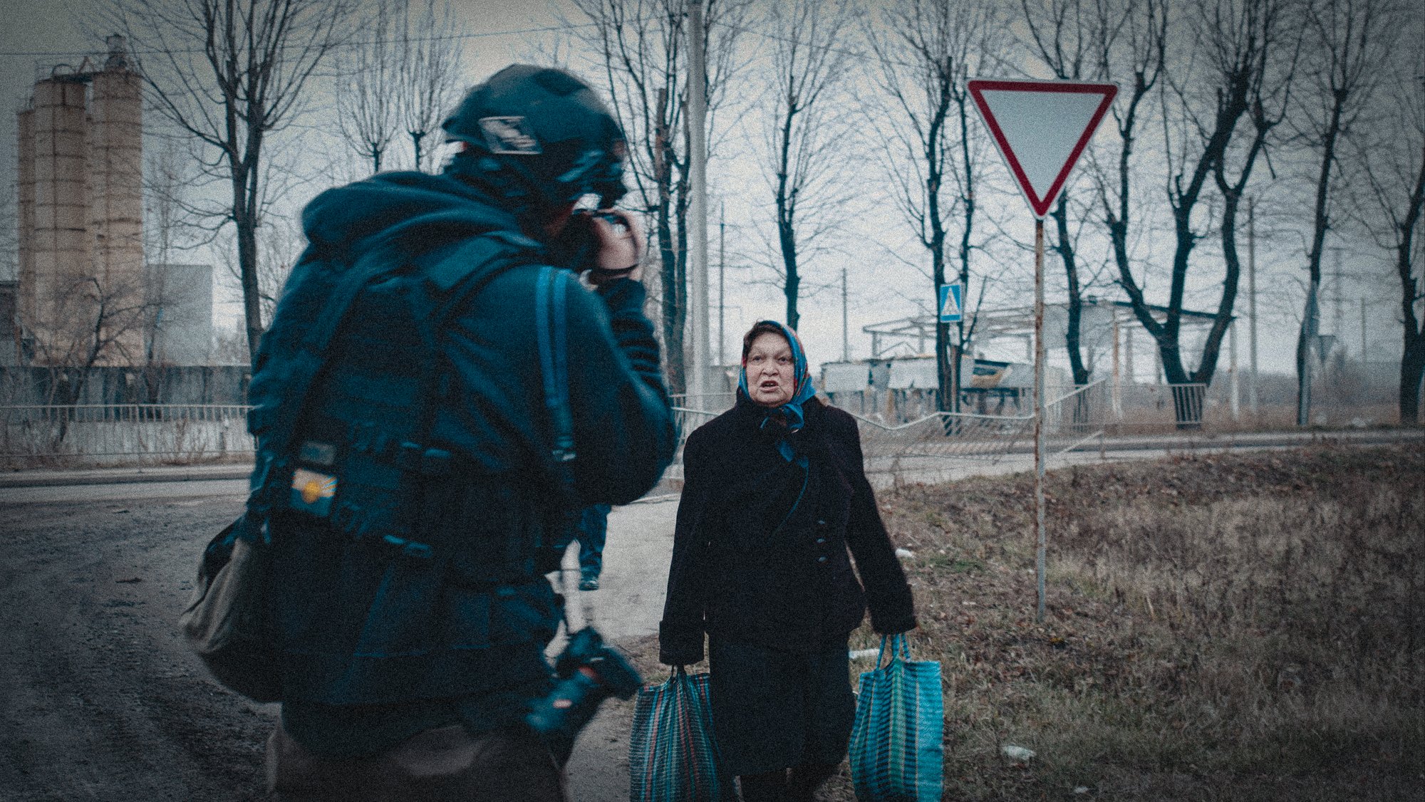 A woman with a headscarf and a man with a helmet are standing having a conversation on the street, with leafless trees and a yield traffic sign in the background.