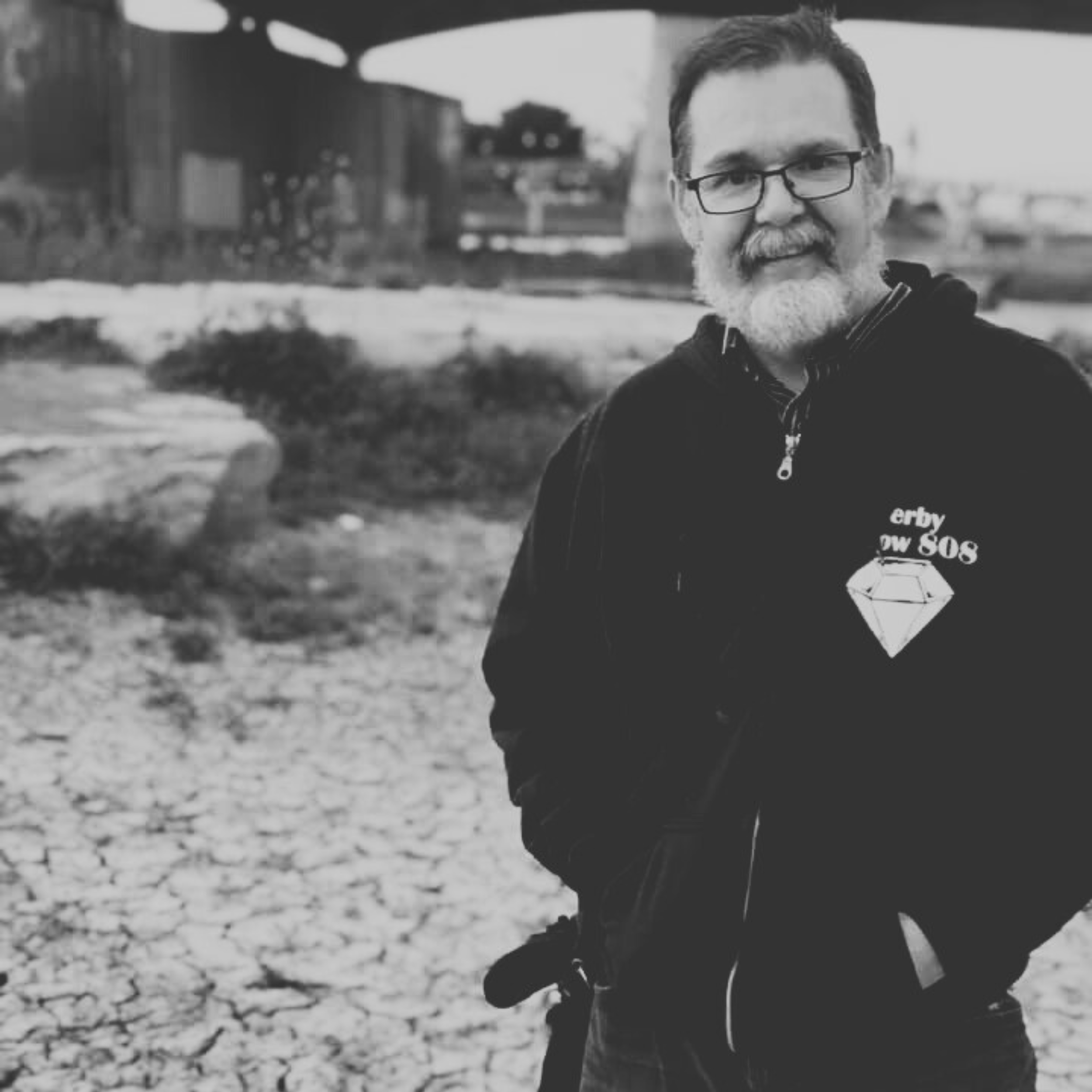 A man with a beard and glasses wearing a dark hoodie stands outdoors in a rocky, industrial area. The background features a large overpass and scattered vegetation. The image is in black and white.