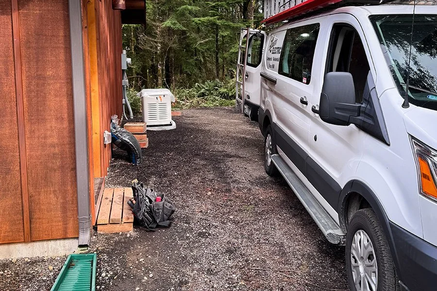White utility van parked on a gravel driveway next to a wooden building, with HVAC equipment and tools visible, surrounded by trees and greenery.