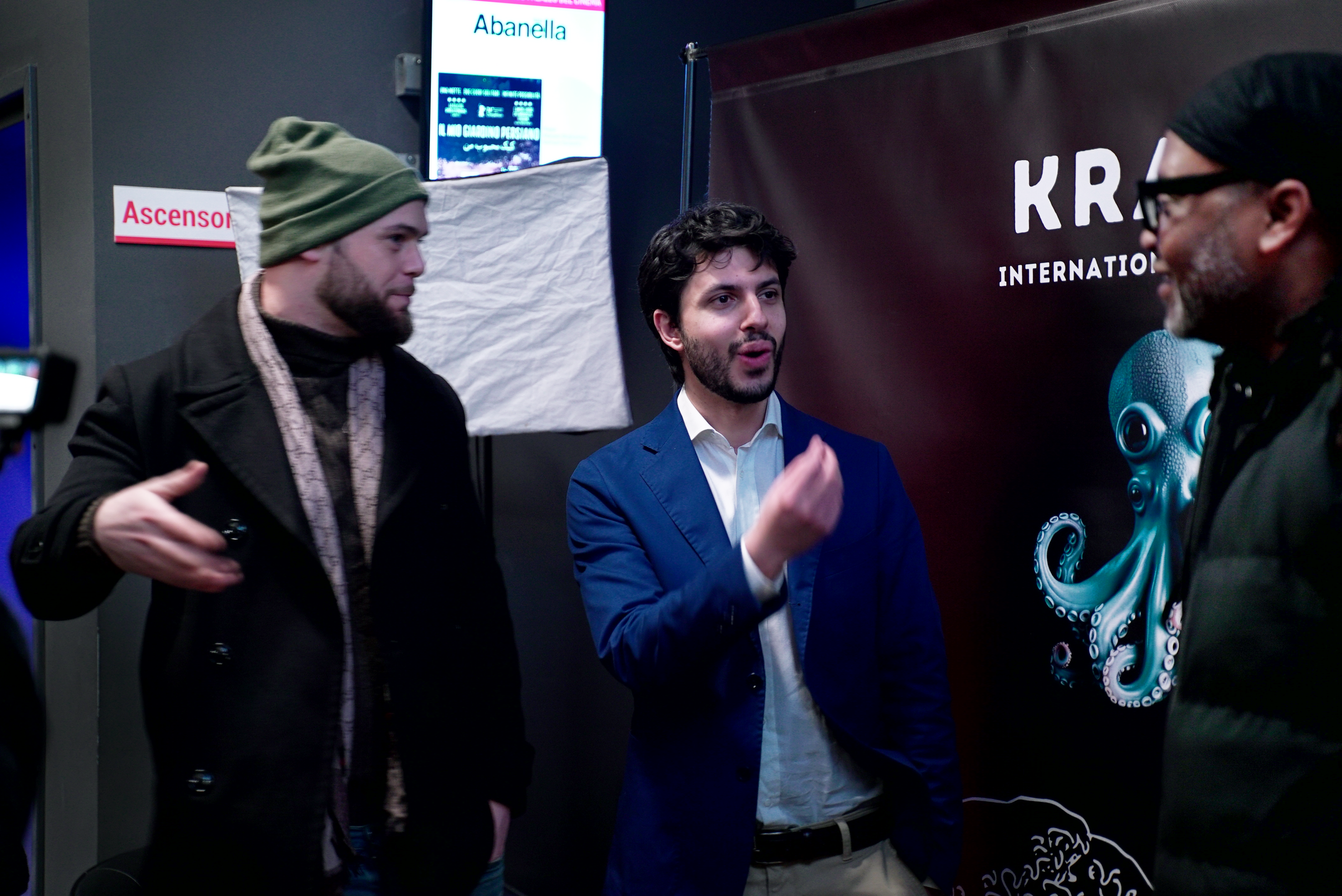 Three men engaged in conversation against a backdrop featuring a colorful octopus illustration and text. One is in a green beanie and black coat, another in a blue blazer, and the third wears glasses. The setting appears casual yet business-oriented.