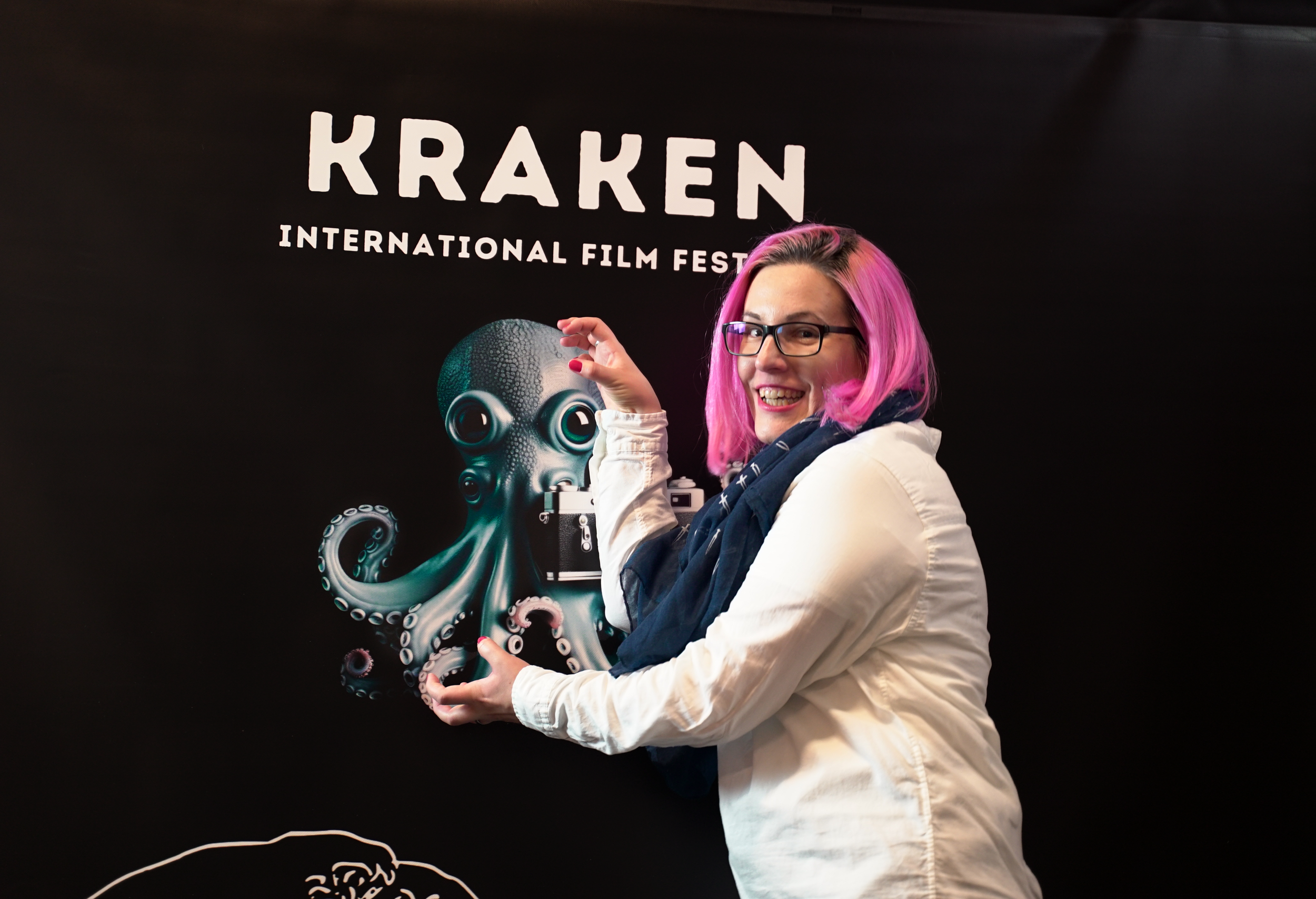 Person with pink hair posing in front of a Kraken International Film Festival banner featuring an octopus illustration.
