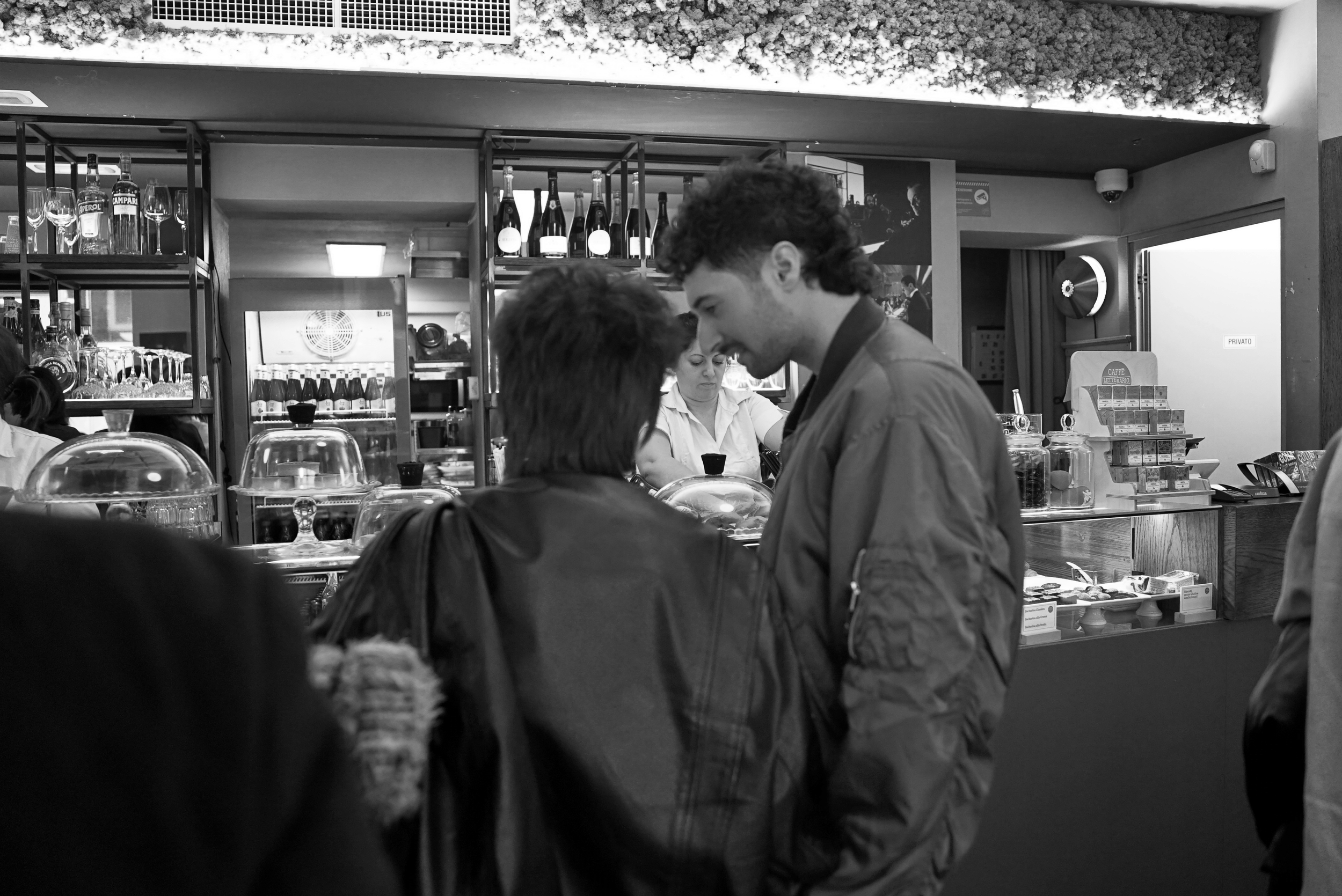 Black and white photo of people at a bar counter with drinks and glassware on shelves, and a bartender in the background.