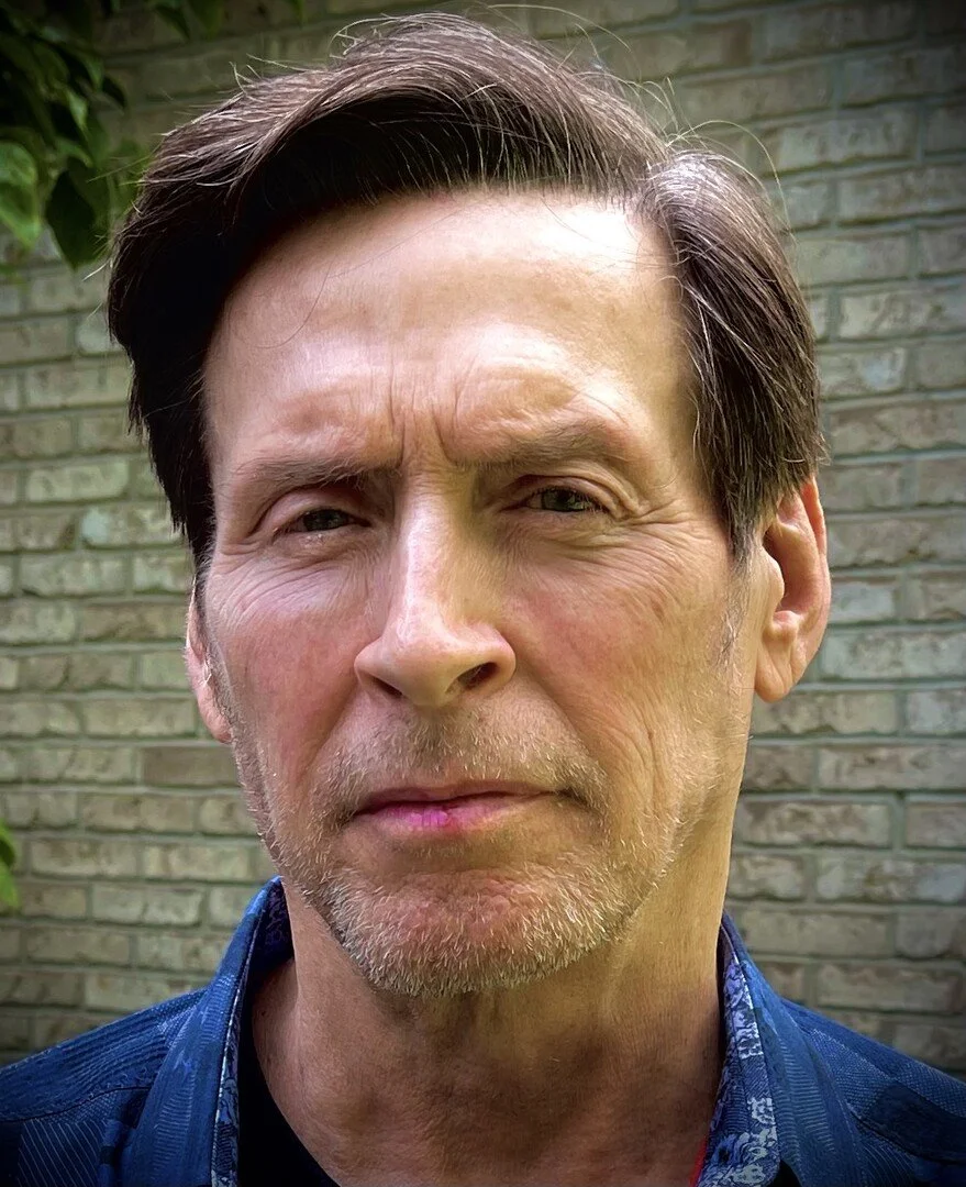 Portrait of an older man with short hair, wearing a dark blue shirt, standing in front of a brick wall.