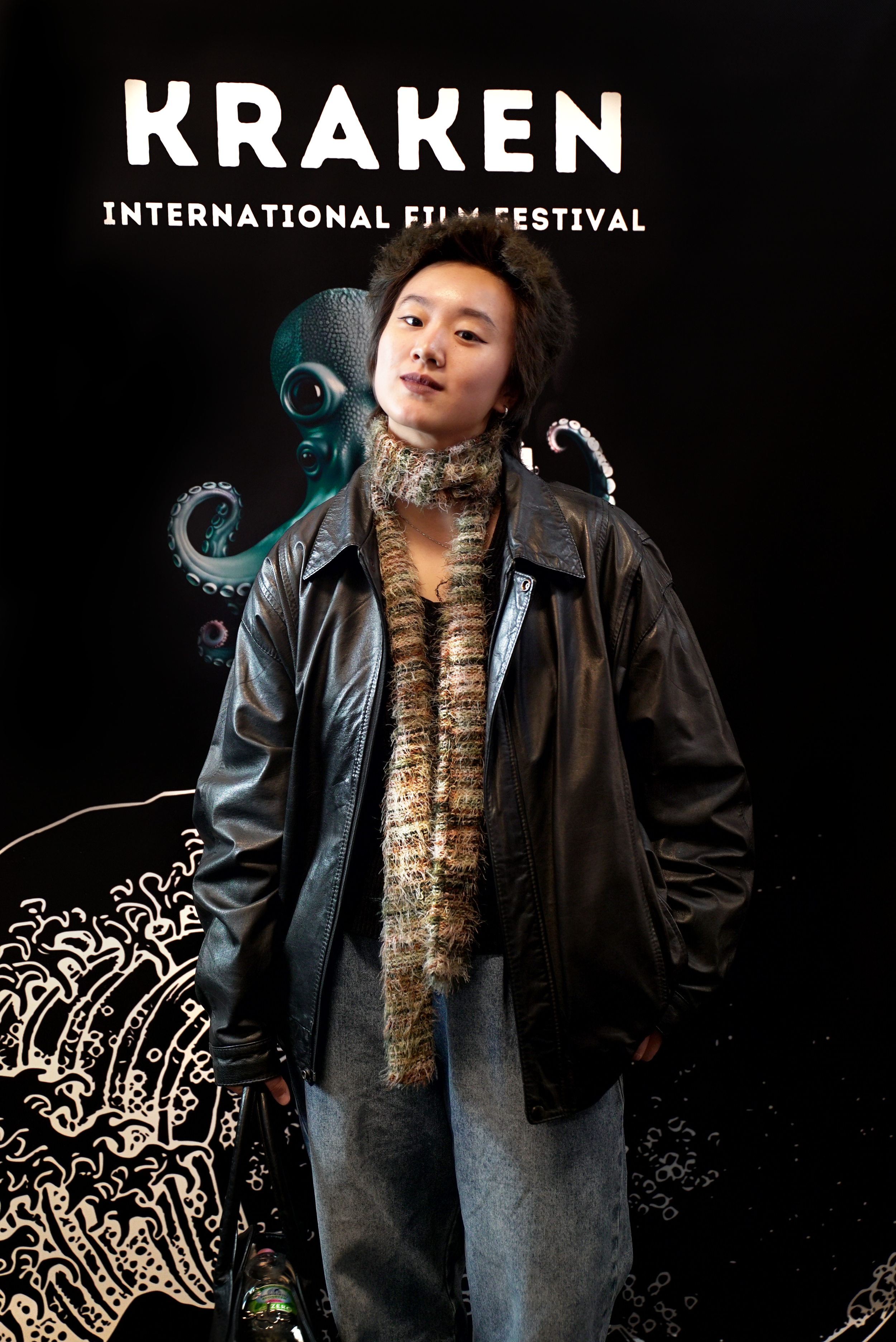 Person in a black leather jacket and knit scarf stands in front of a backdrop for the Kraken International Film Festival, featuring a kraken illustration.