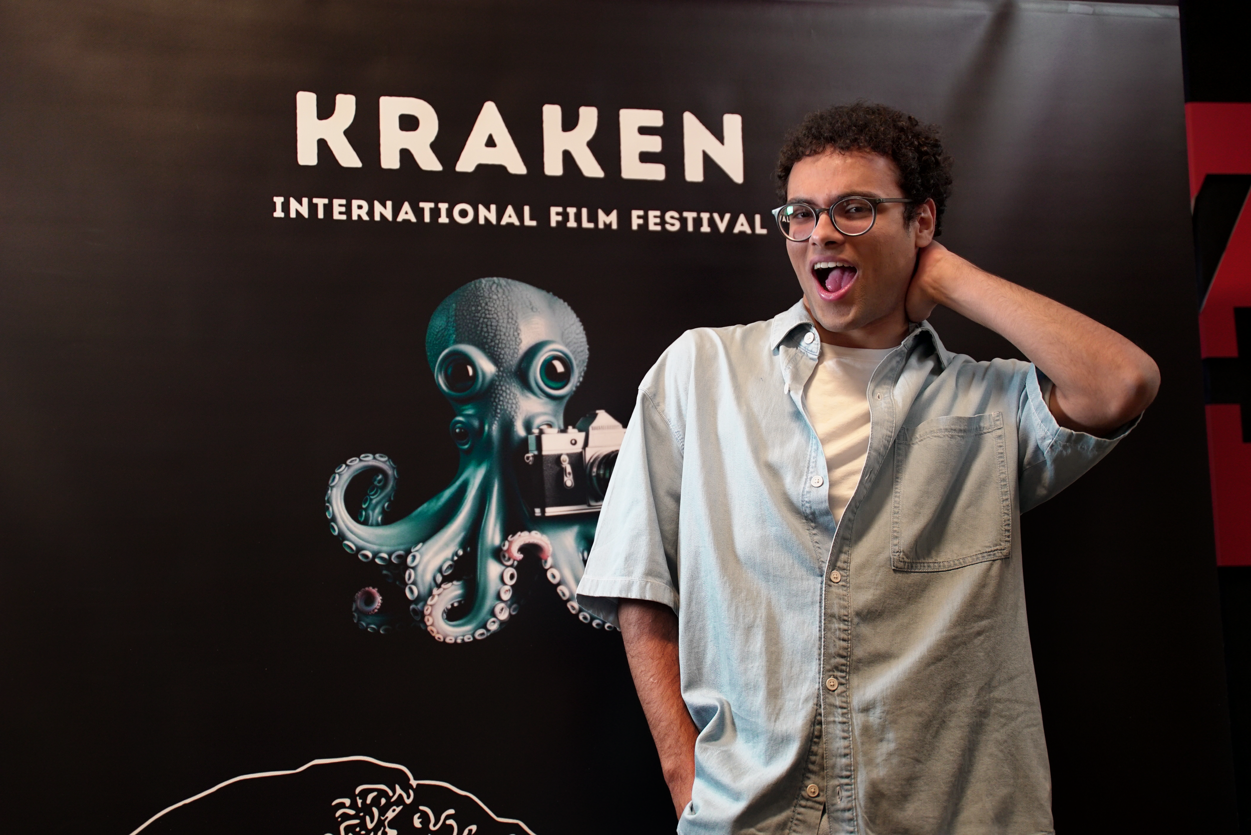 Person posing playfully in front of a "Kraken International Film Festival" banner featuring an octopus with a camera.