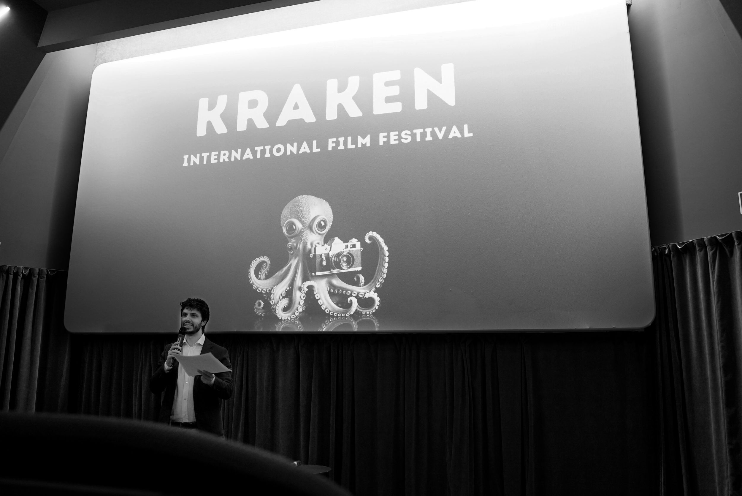 Person speaking into a microphone in front of a screen displaying "KRAKEN International Film Festival" with an octopus holding a camera illustration.