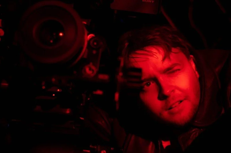 A man illuminated by red light looking at the camera beside a large camera lens, with a squinting expression.