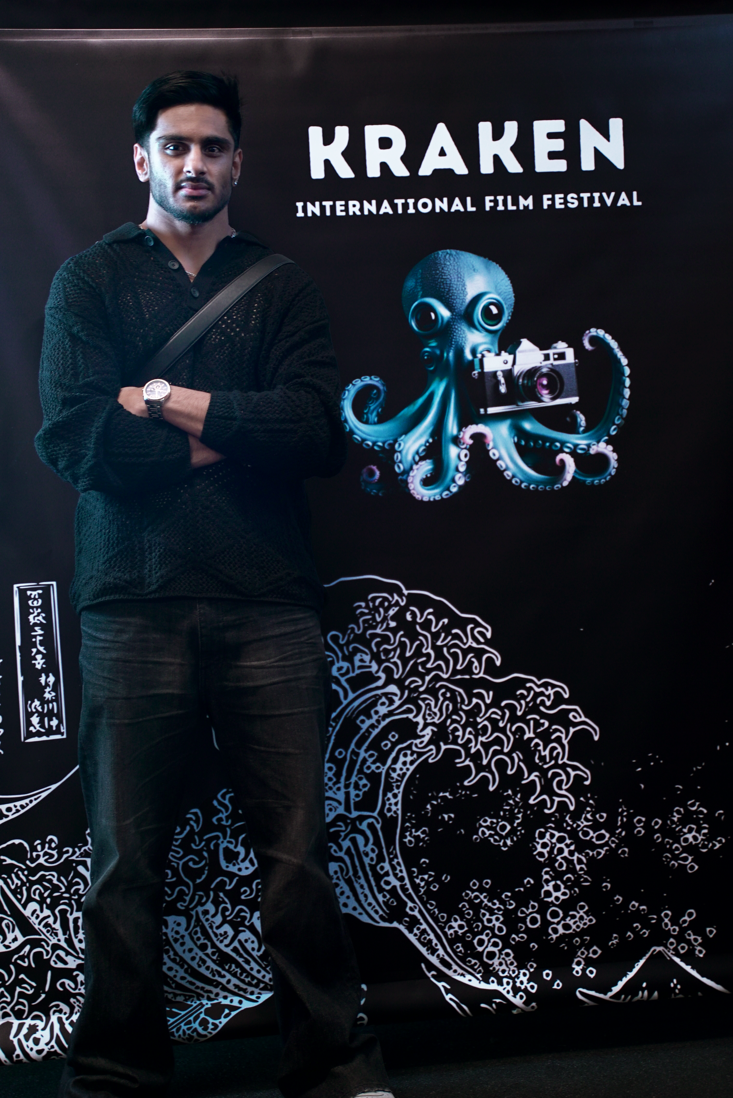 A person standing in front of a banner for the Kraken International Film Festival. The banner features an illustrated octopus holding a camera and a stylized wave design.